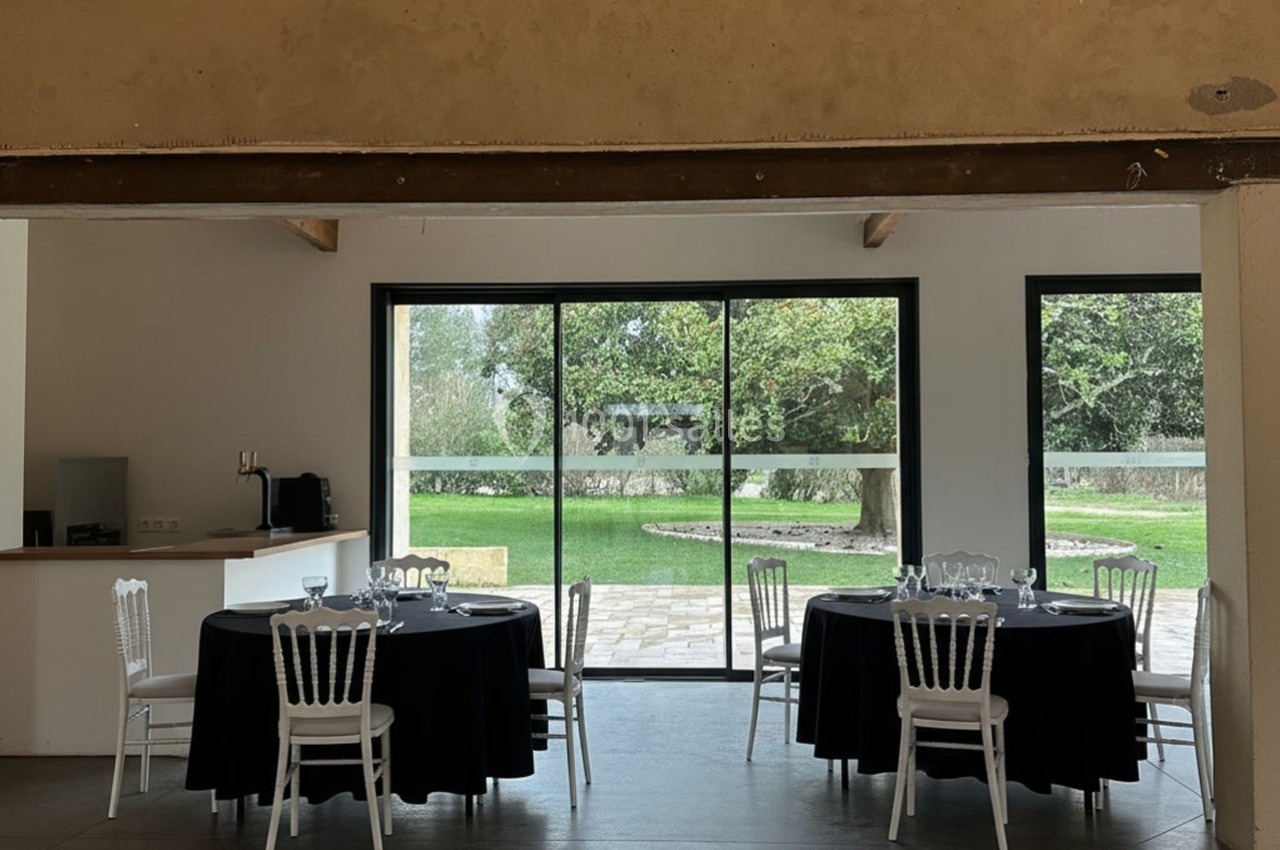 Salle lumineuse avec tables rondes dressées, nappes noires, chaises blanches et vue sur un jardin verdoyant par de grandes…