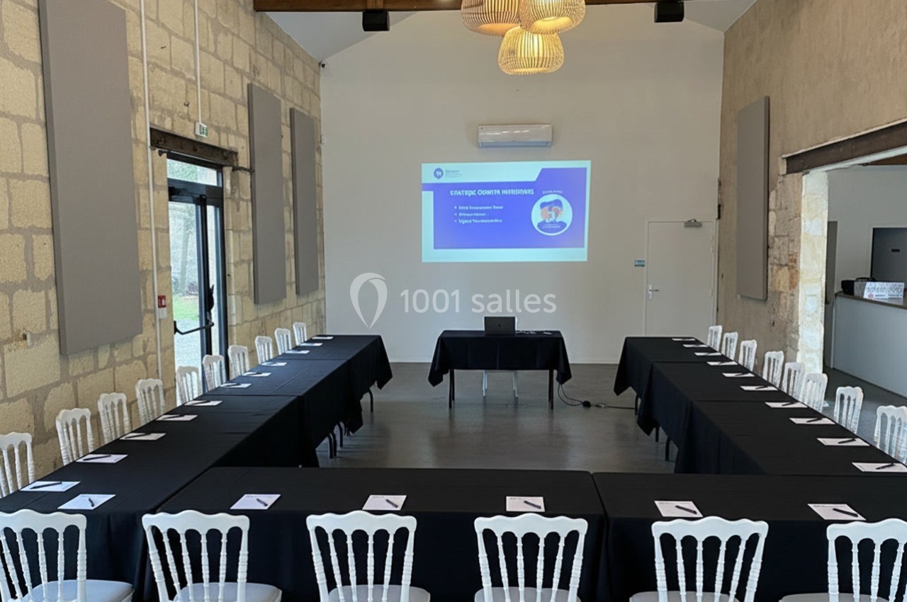 Salle de réunion lumineuse avec tables en U, chaises blanches et écran de projection affichant une présentation.