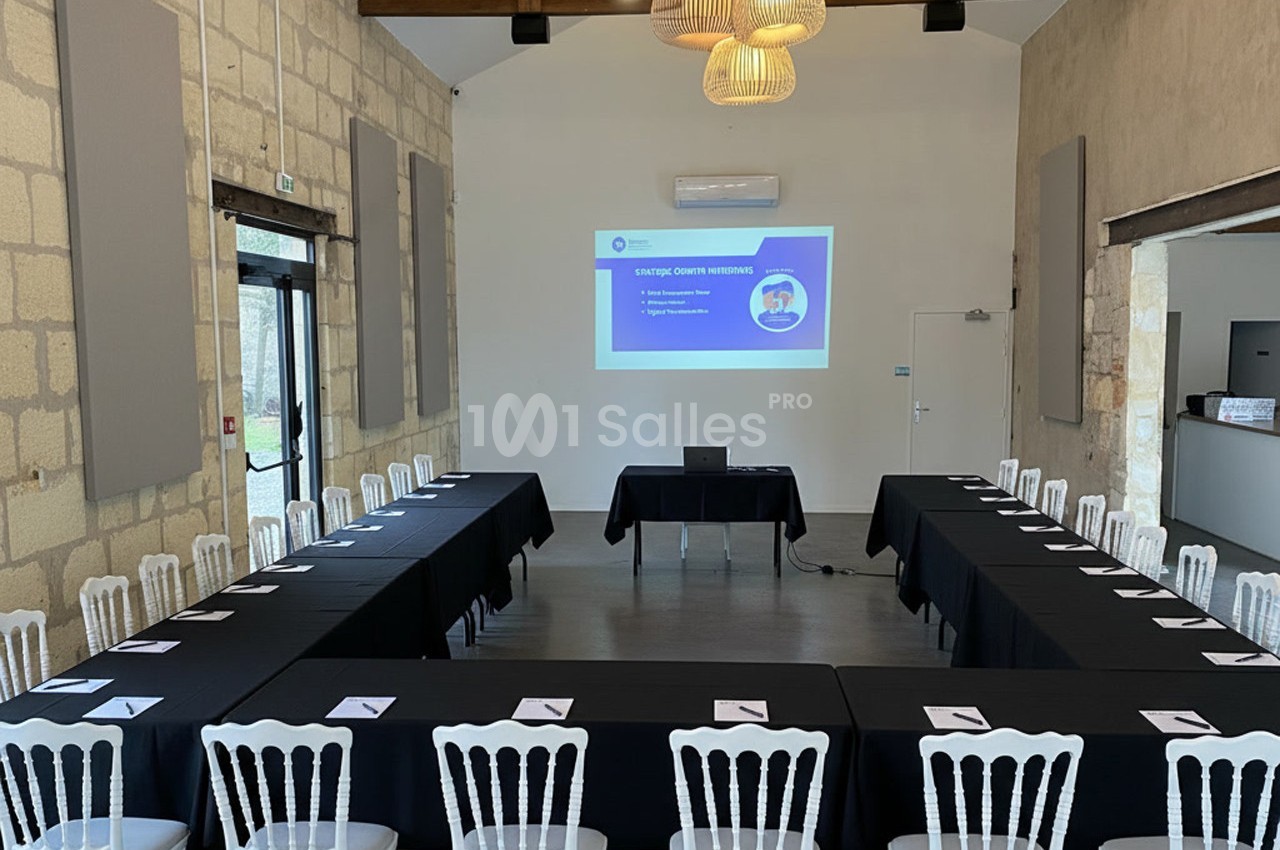 Salle de réunion lumineuse avec tables en U, chaises blanches et écran de projection affichant une présentation.