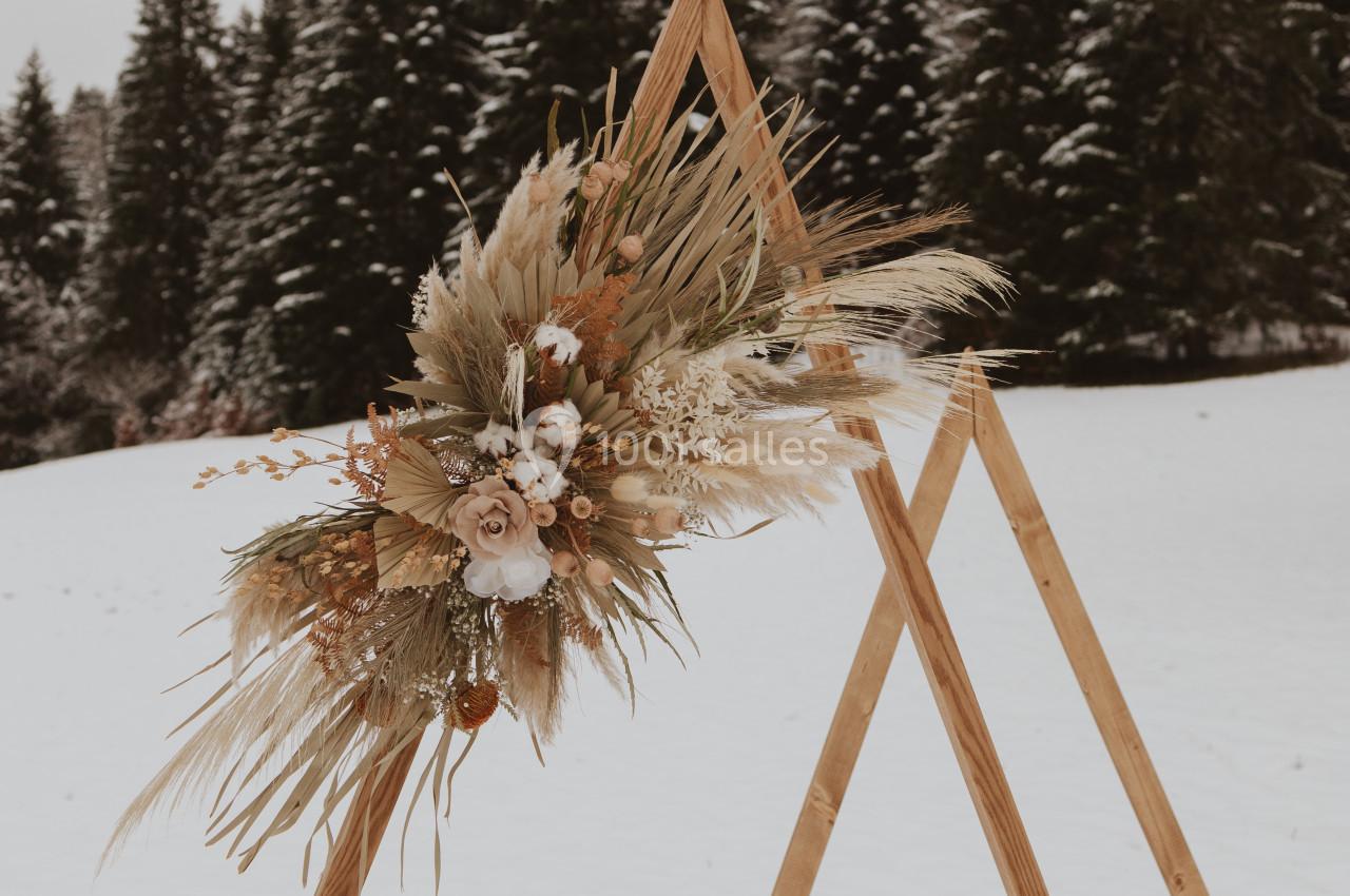 Structure en bois triangulaire décorée de fleurs séchées et de pampas, placée dans un paysage enneigé avec des sapins.