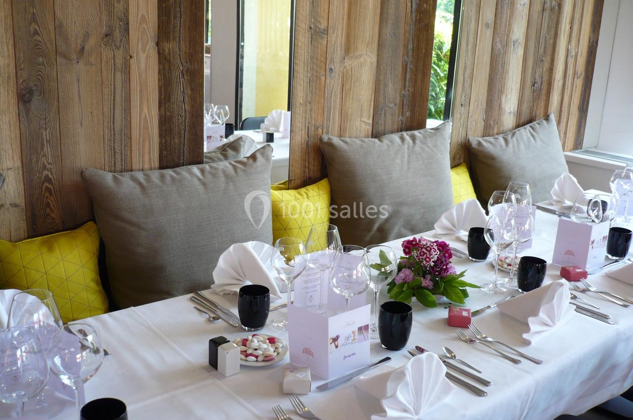 Table dressée avec nappes blanches, serviettes pliées, verres, fleurs et coussins jaunes sur une banquette en bois.
