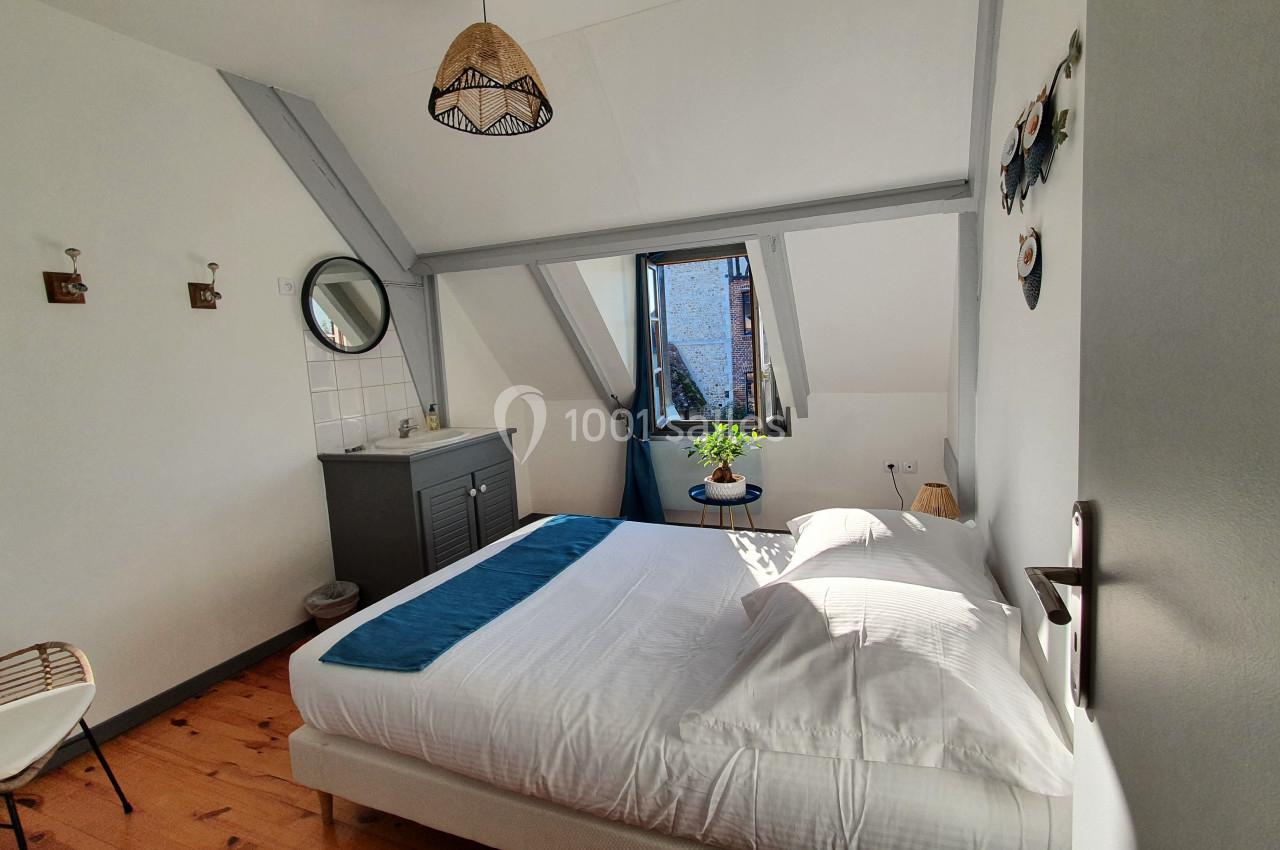 Chambre lumineuse sous combles avec lit double, commode, miroir rond, fenêtre ouverte et décoration murale.