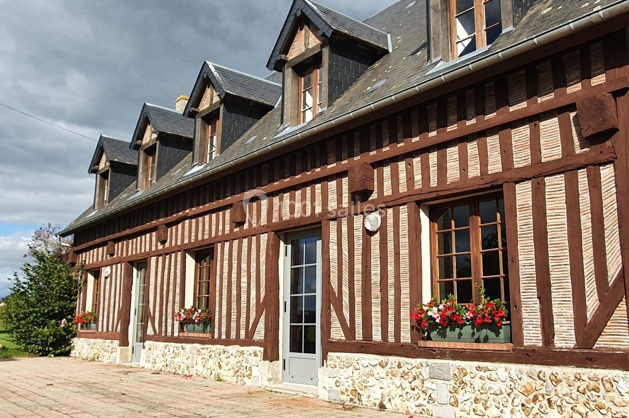 Façade d'une maison à colombages avec fenêtres fleuries, toit en ardoise et ciel partiellement nuageux.