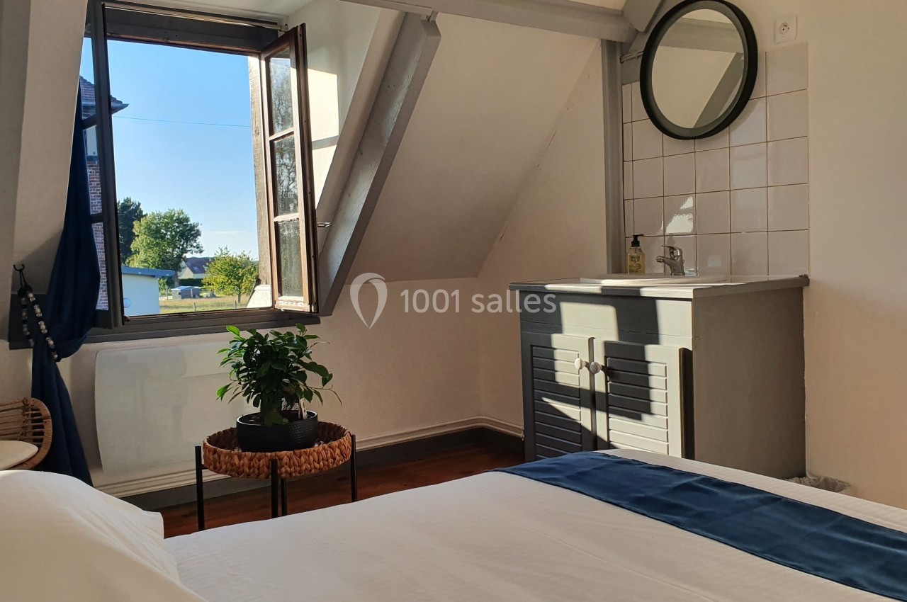 Chambre lumineuse avec lit, fenêtre ouverte sur un paysage, lavabo avec miroir et plante sur une table basse.
