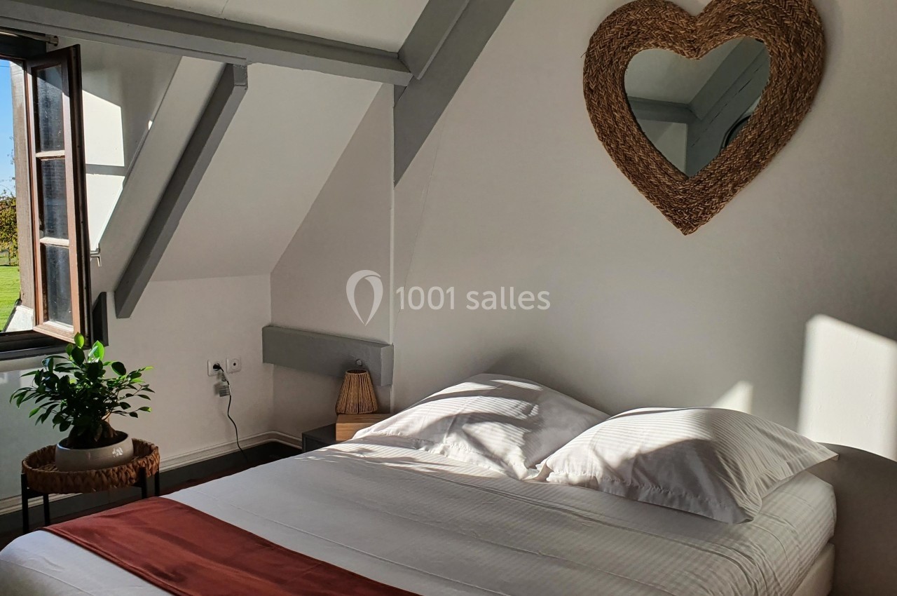 Chambre lumineuse avec un lit double, un miroir en forme de cœur au mur et une fenêtre ouverte donnant sur un jardin.