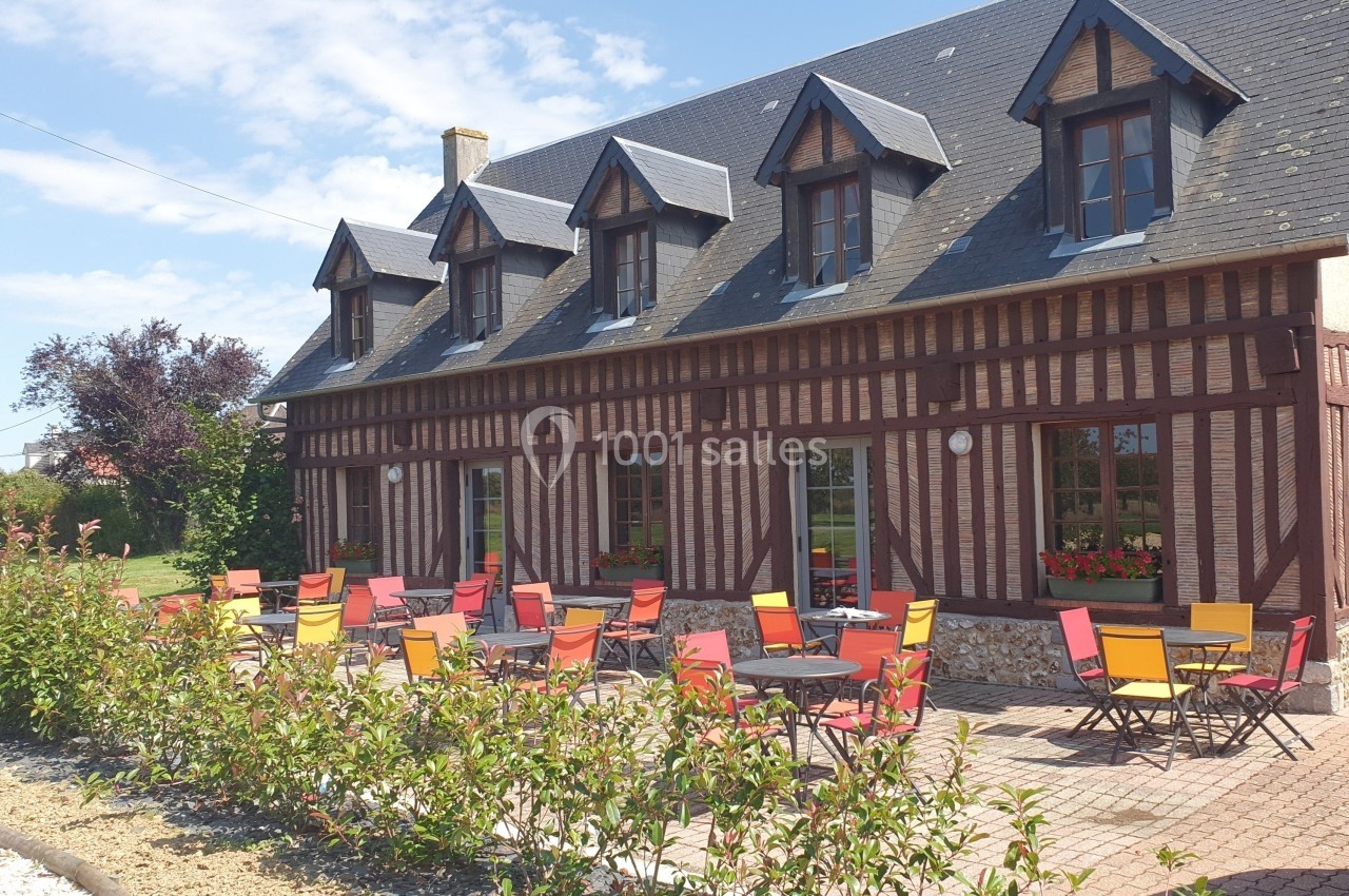 Façade d'une maison à colombages avec terrasse aménagée de tables et chaises colorées, sous un ciel dégagé.