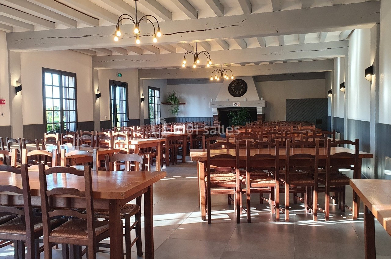 Salle de restaurant lumineuse avec des tables et chaises en bois, grandes fenêtres et une cheminée en fond.