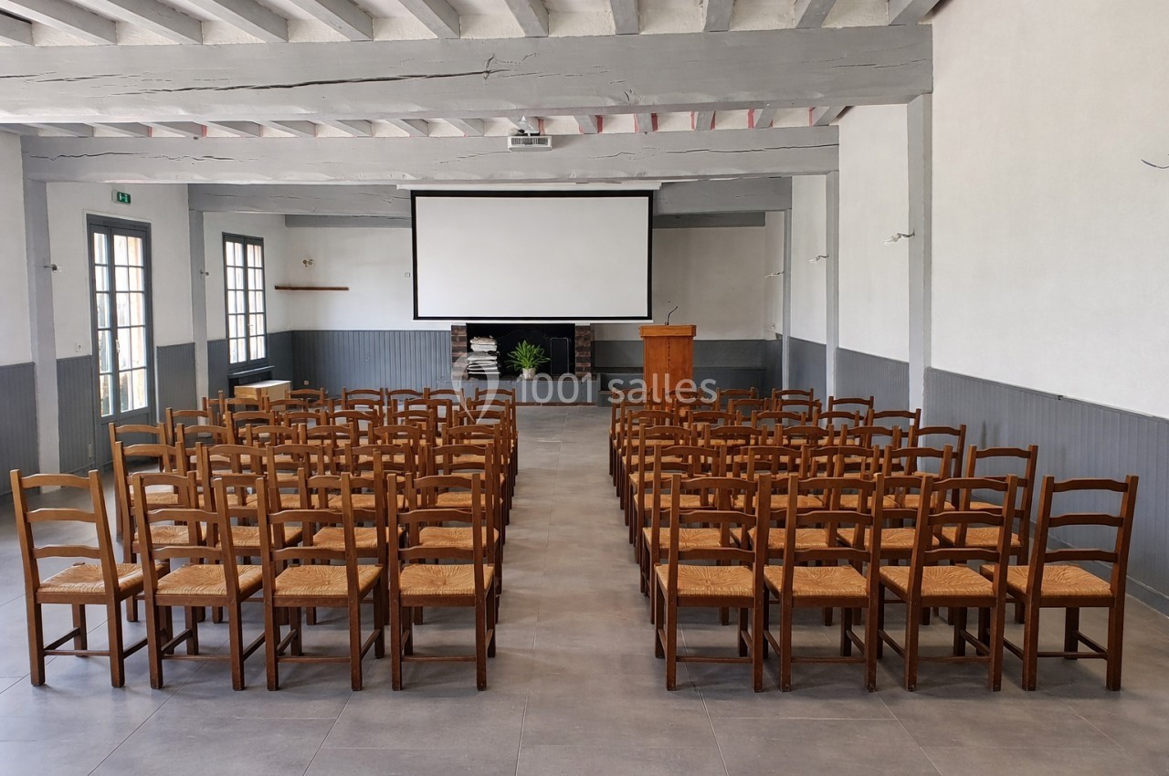 Salle avec rangées de chaises en bois, écran de projection, pupitre en bois et murs clairs.