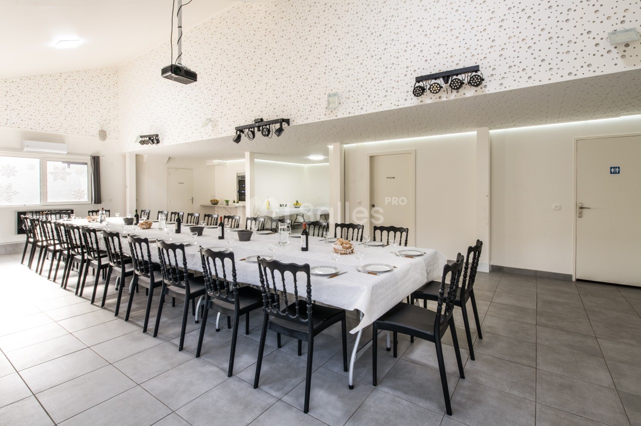 Grande salle lumineuse avec deux longues tables dressées, chaises noires, murs blancs et décor minimaliste.