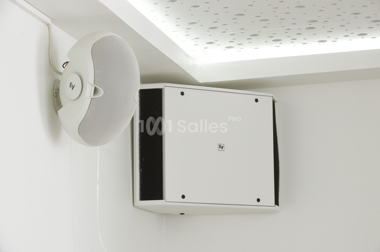 Haut-parleur mural blanc fixé dans un angle, avec un plafond perforé visible en arrière-plan.