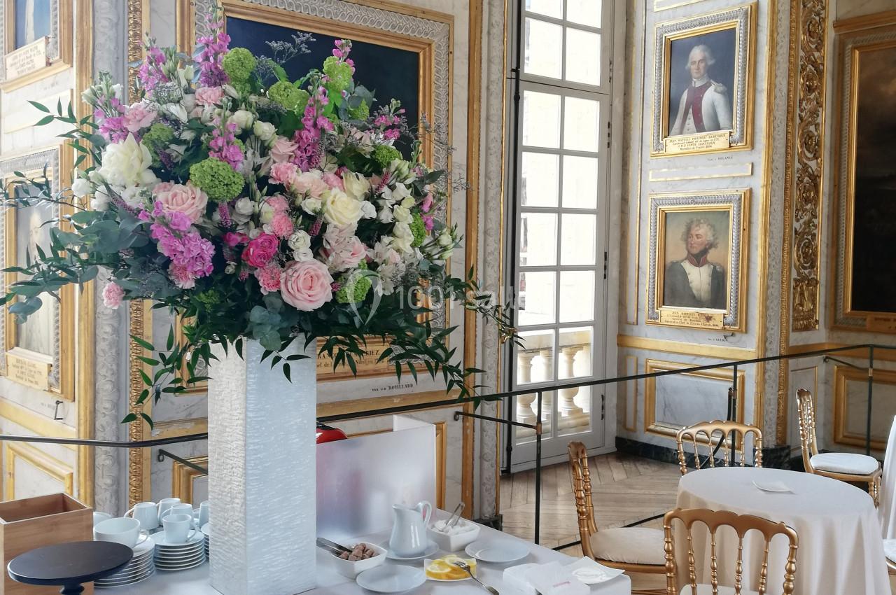 Grande salle ornée de portraits et dorures, avec une table dressée, décorée d'un grand bouquet de fleurs colorées.