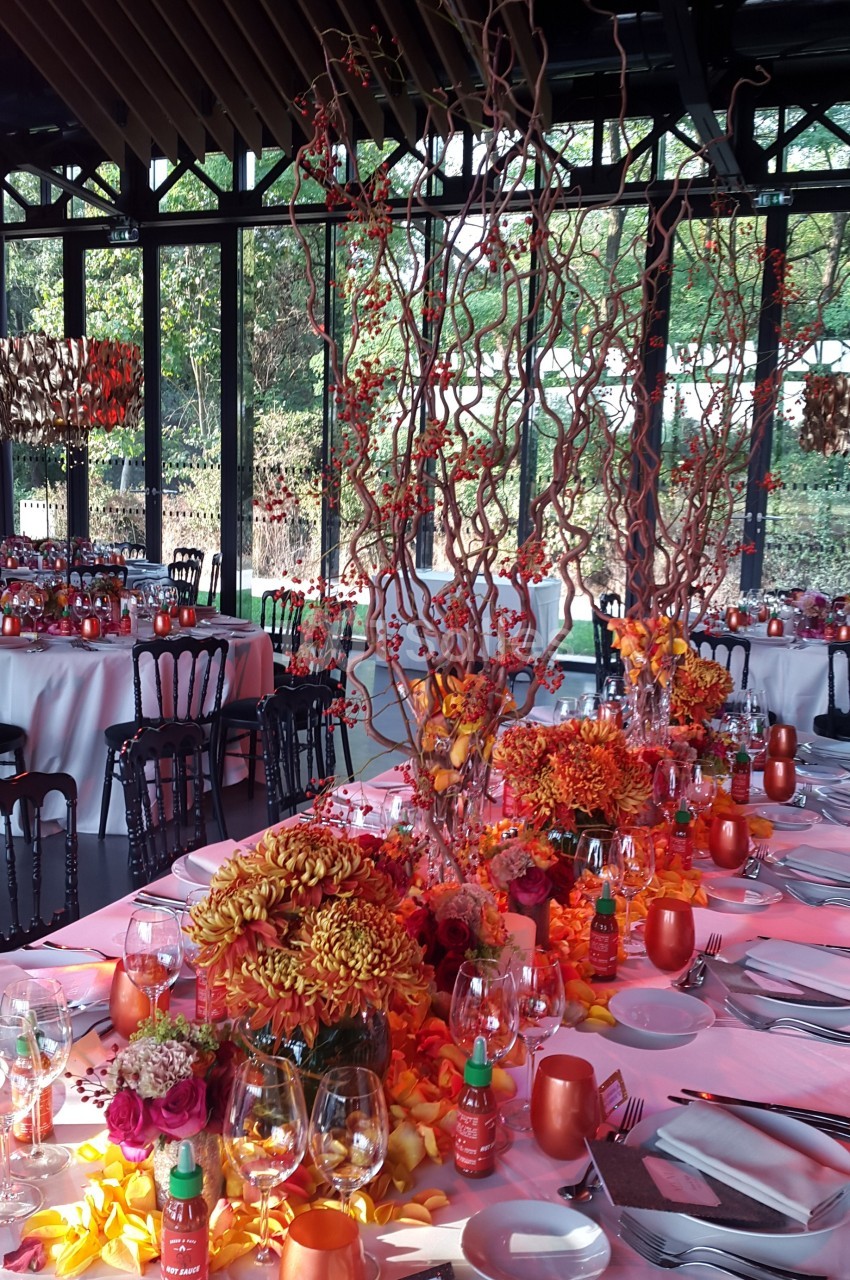 Table décorée pour un événement avec des fleurs orange, branches torsadées et vaisselle élégante dans une salle vitrée.