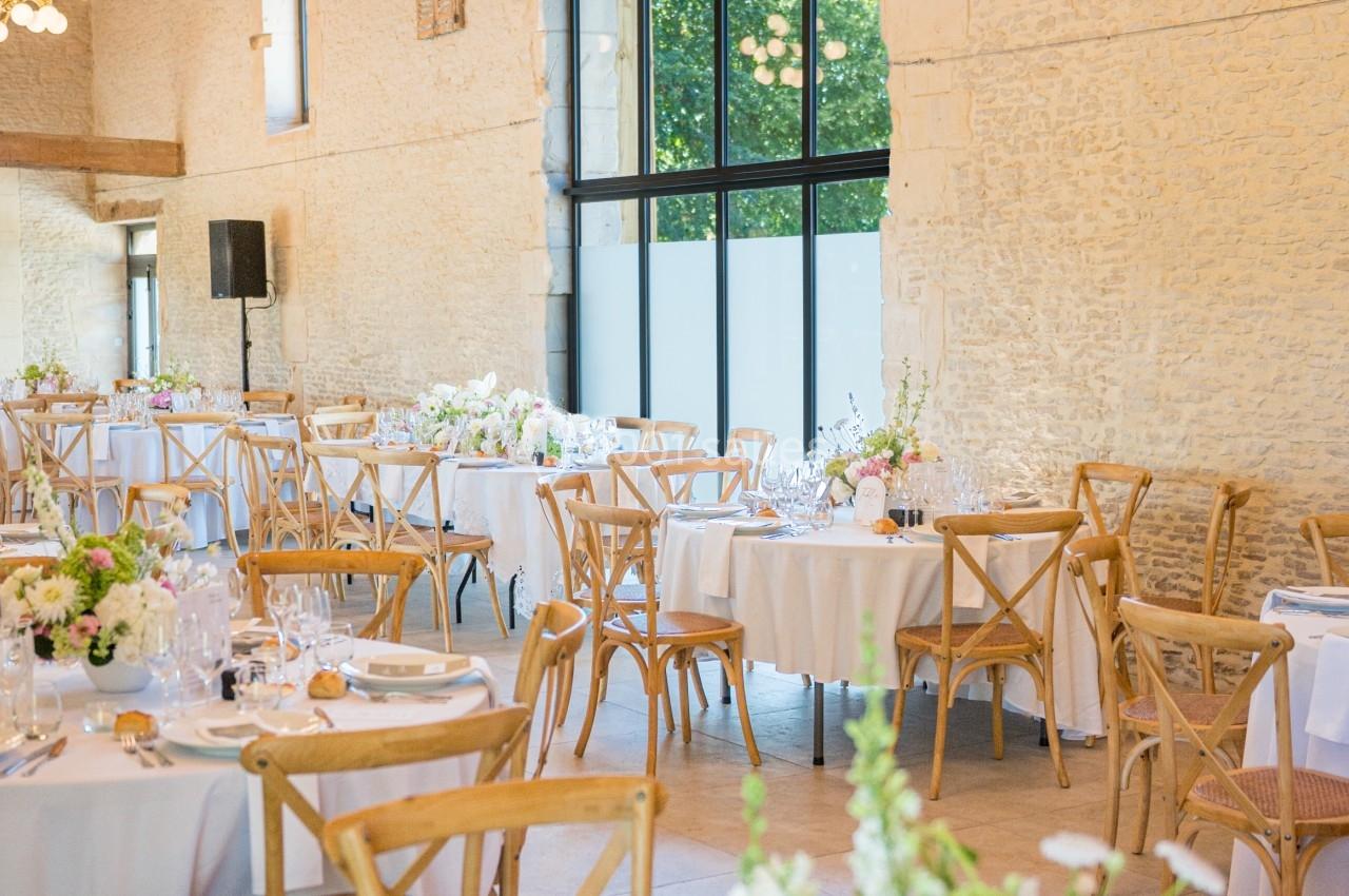 Salle de réception lumineuse avec tables décorées de fleurs, chaises en bois et grandes fenêtres donnant sur l'extérieur.