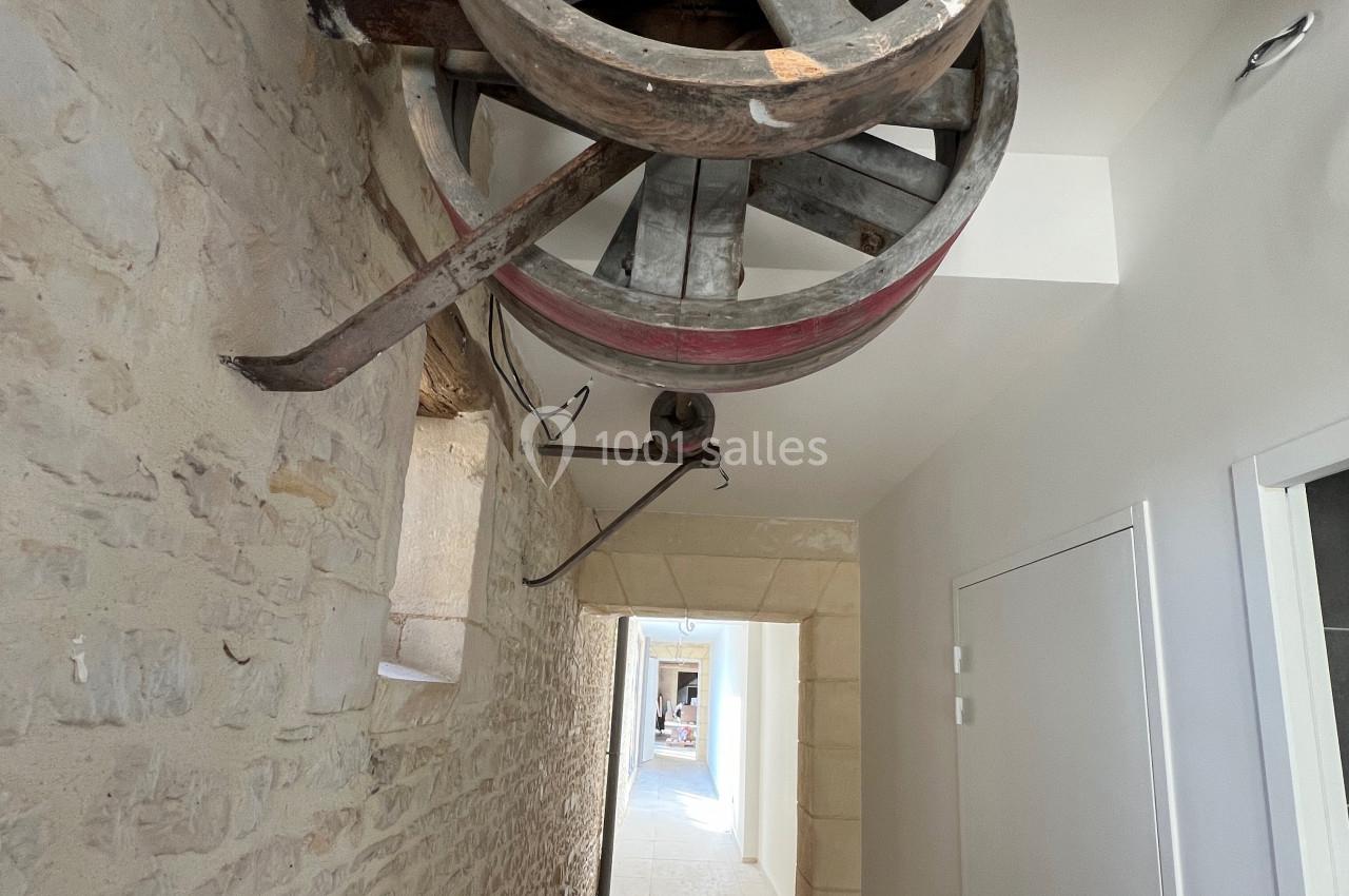 Roue en bois suspendue au plafond d'un couloir aux murs en pierre, avec une porte et un passage lumineux au fond.
