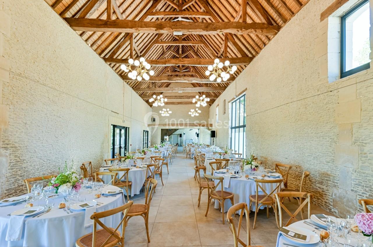 Salle de réception lumineuse avec tables dressées, chaises en bois et poutres apparentes sous un plafond haut.