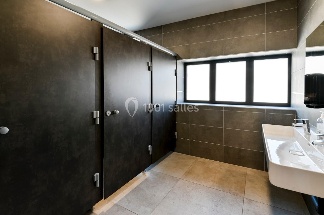 Cabines de toilettes modernes avec portes sombres, lavabo mural blanc et grande fenêtre laissant entrer la lumière naturelle.