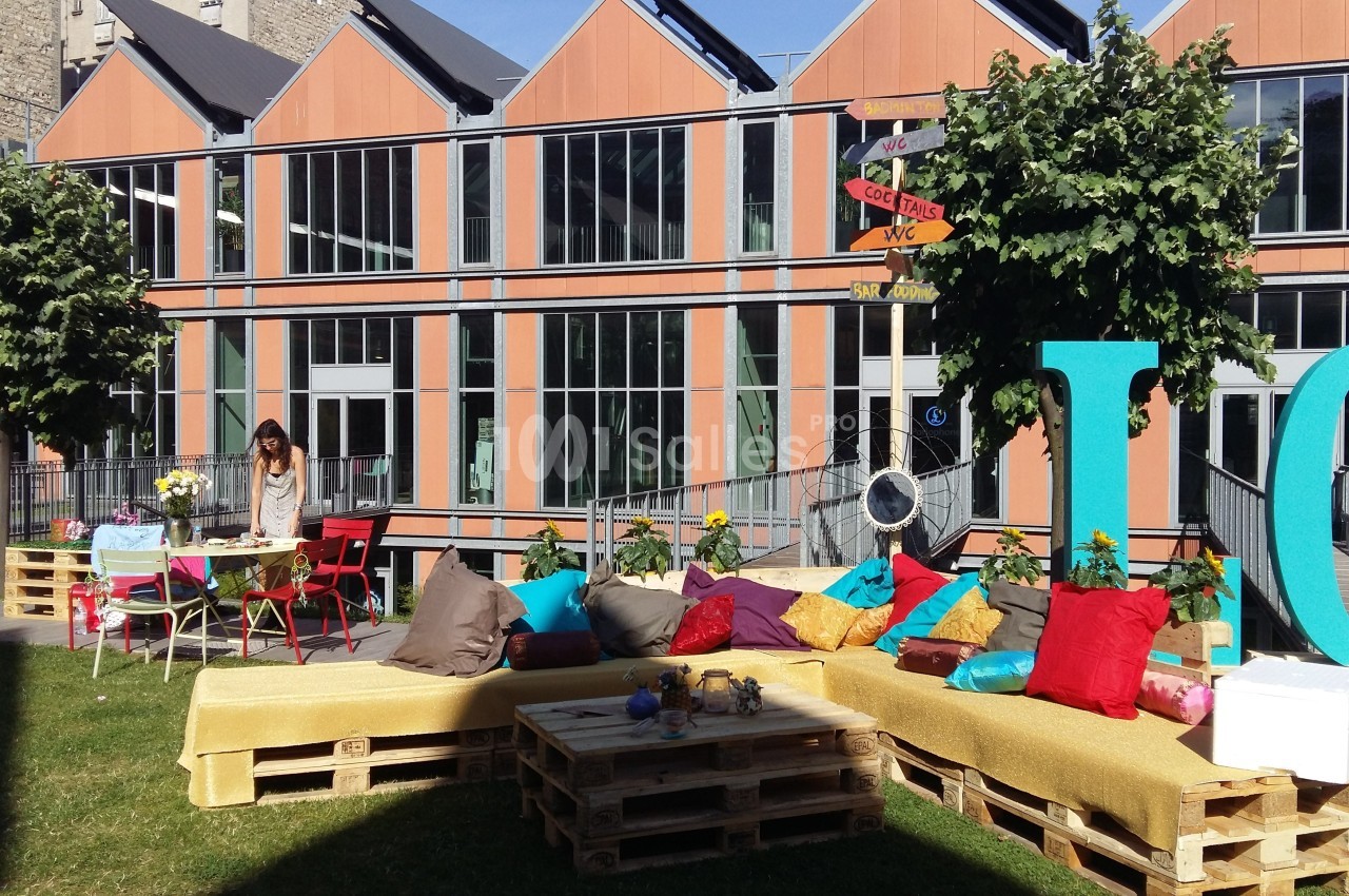 Espace extérieur aménagé avec des palettes, coussins colorés et tables, devant un bâtiment moderne avec grandes fenêtres.