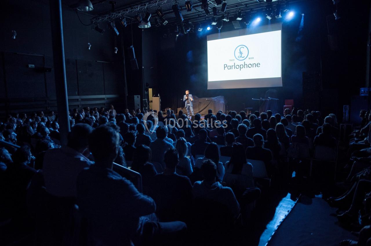 Salle de spectacle sombre avec un public assis face à une scène éclairée où une personne parle devant un écran.