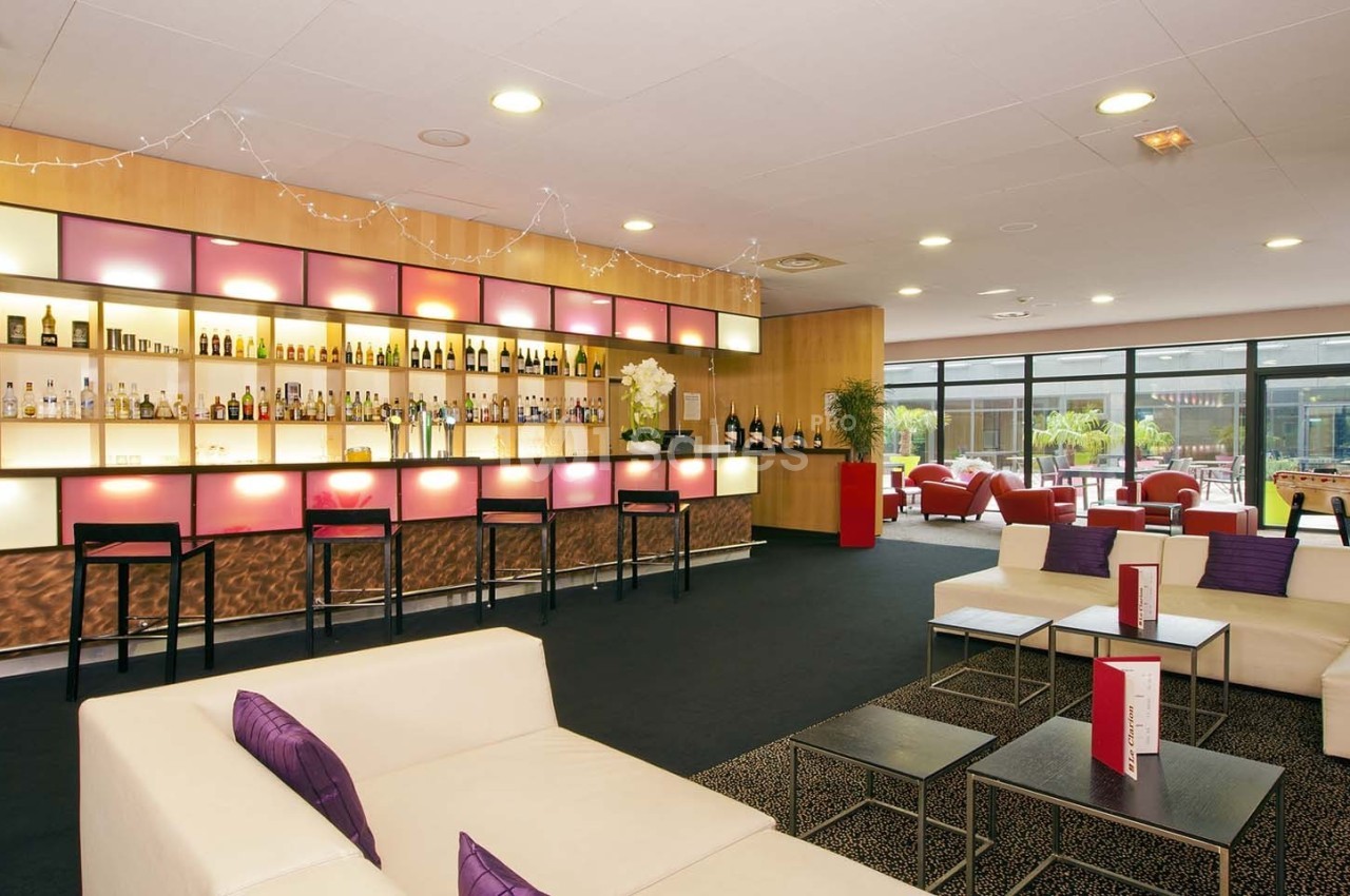 Bar moderne avec étagères illuminées, comptoir avec tabourets, canapés clairs et tables basses dans un espace lumineux.
