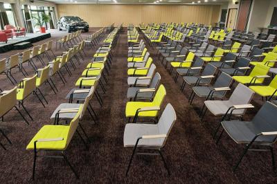 Salle de conférence avec des rangées de chaises grises et jaunes, une scène et une voiture en arrière-plan.