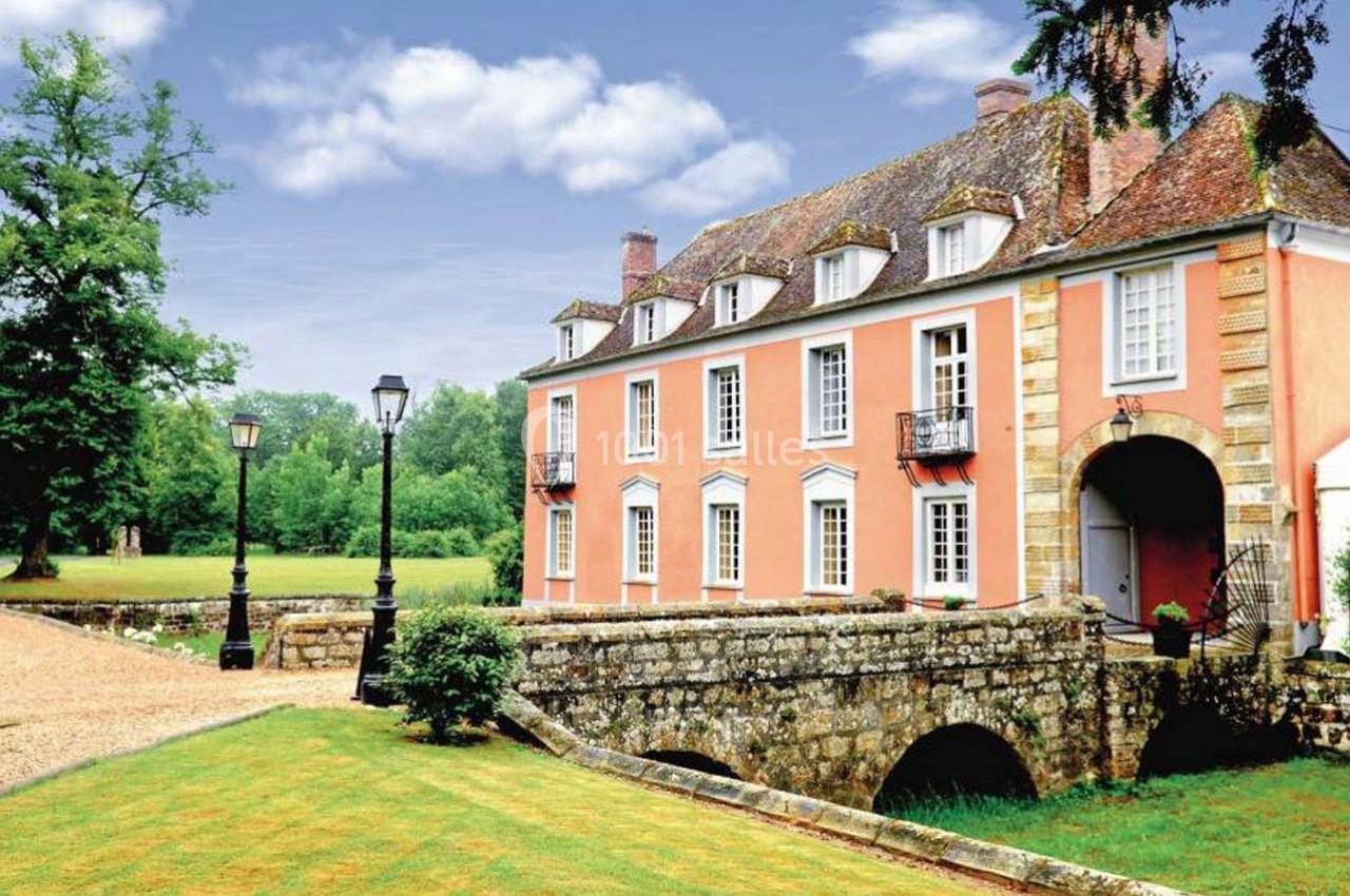Manoir ancien en pierre avec façade rose, entouré de verdure, traversé par un petit pont en arc.