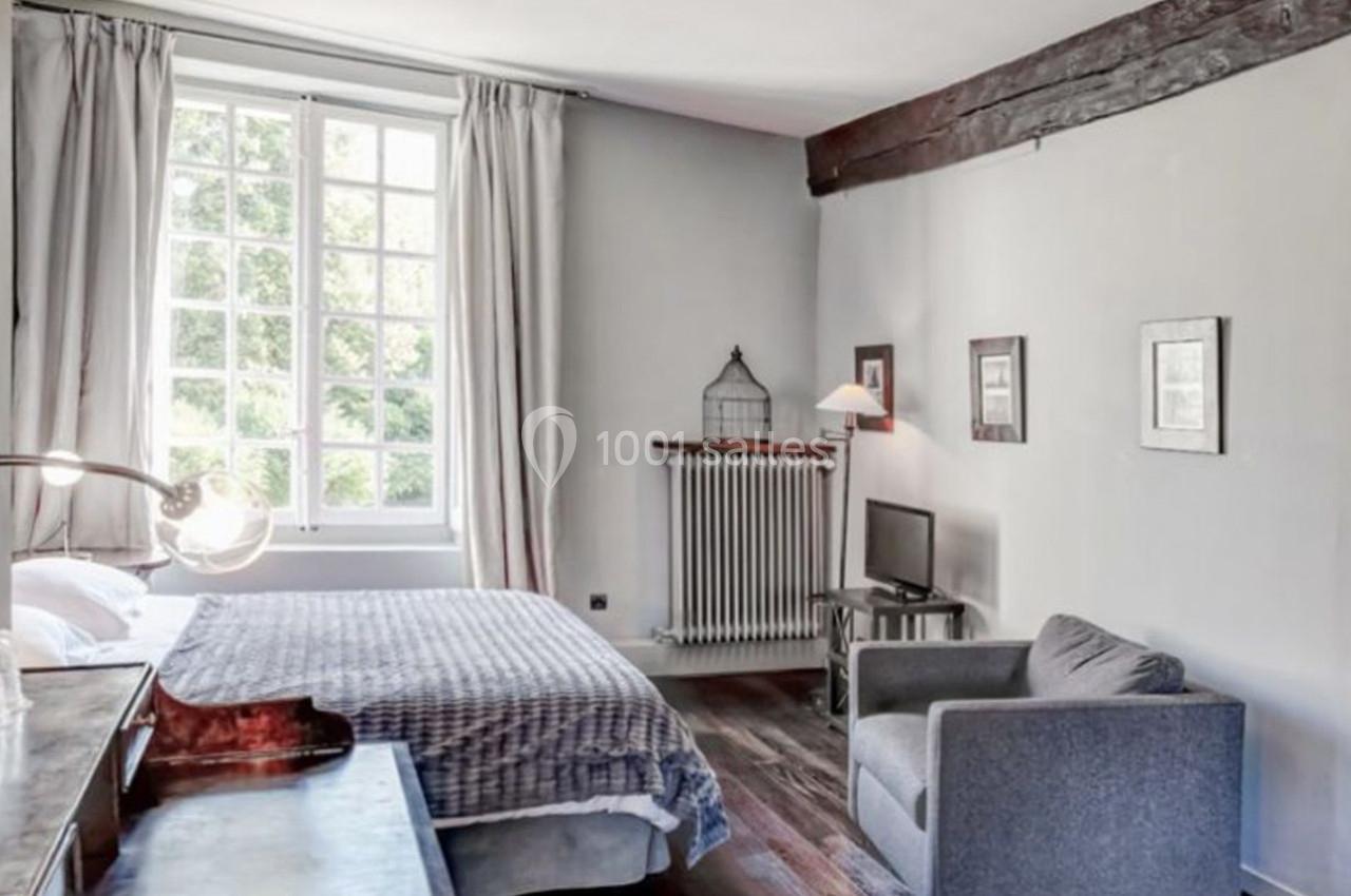 Chambre lumineuse avec lit double, fauteuil gris, bureau en bois, fenêtre à carreaux et poutres apparentes.