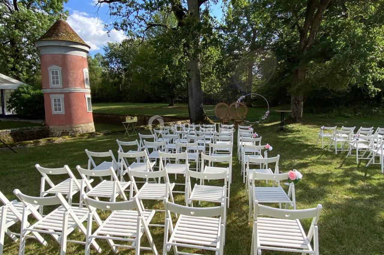 Chaises blanches disposées en rangées sur une pelouse, face à une arche décorative, près d'un petit bâtiment rose.