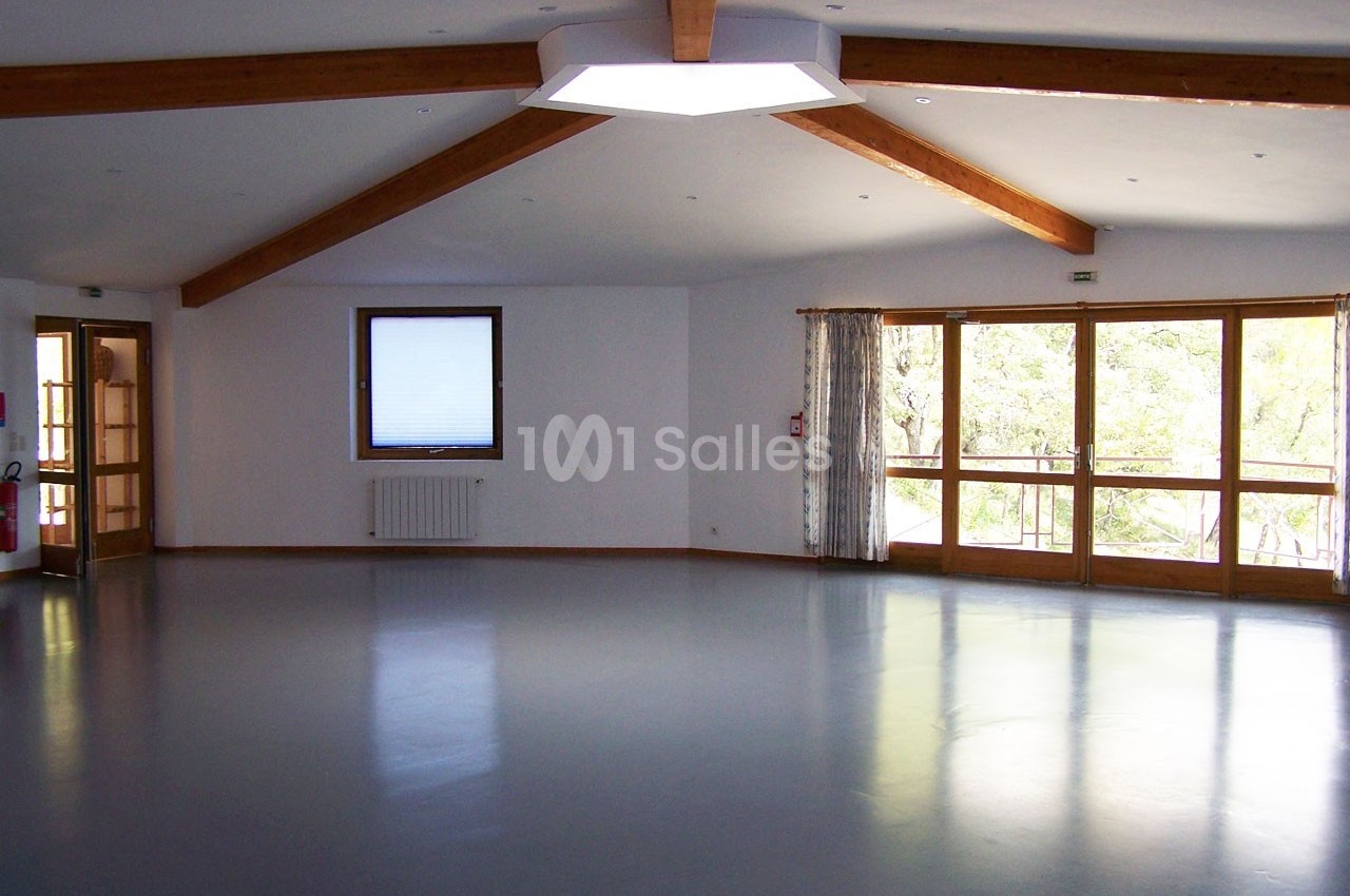 Salle vide avec sol gris, poutres apparentes au plafond, fenêtres laissant entrer la lumière naturelle.