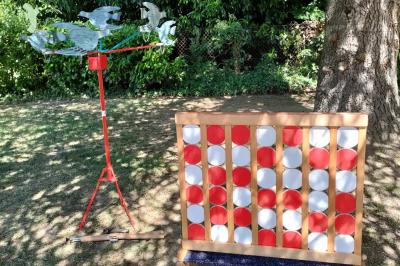 Un jeu de puissance 4 géant en bois placé sur une pelouse, à côté d'une girouette rouge sous des arbres.
