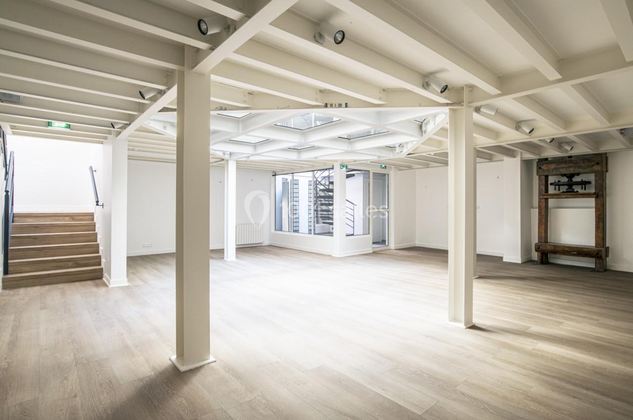 Salle lumineuse avec parquet clair, poutres apparentes blanches, escalier sur la gauche et grande verrière au plafond.