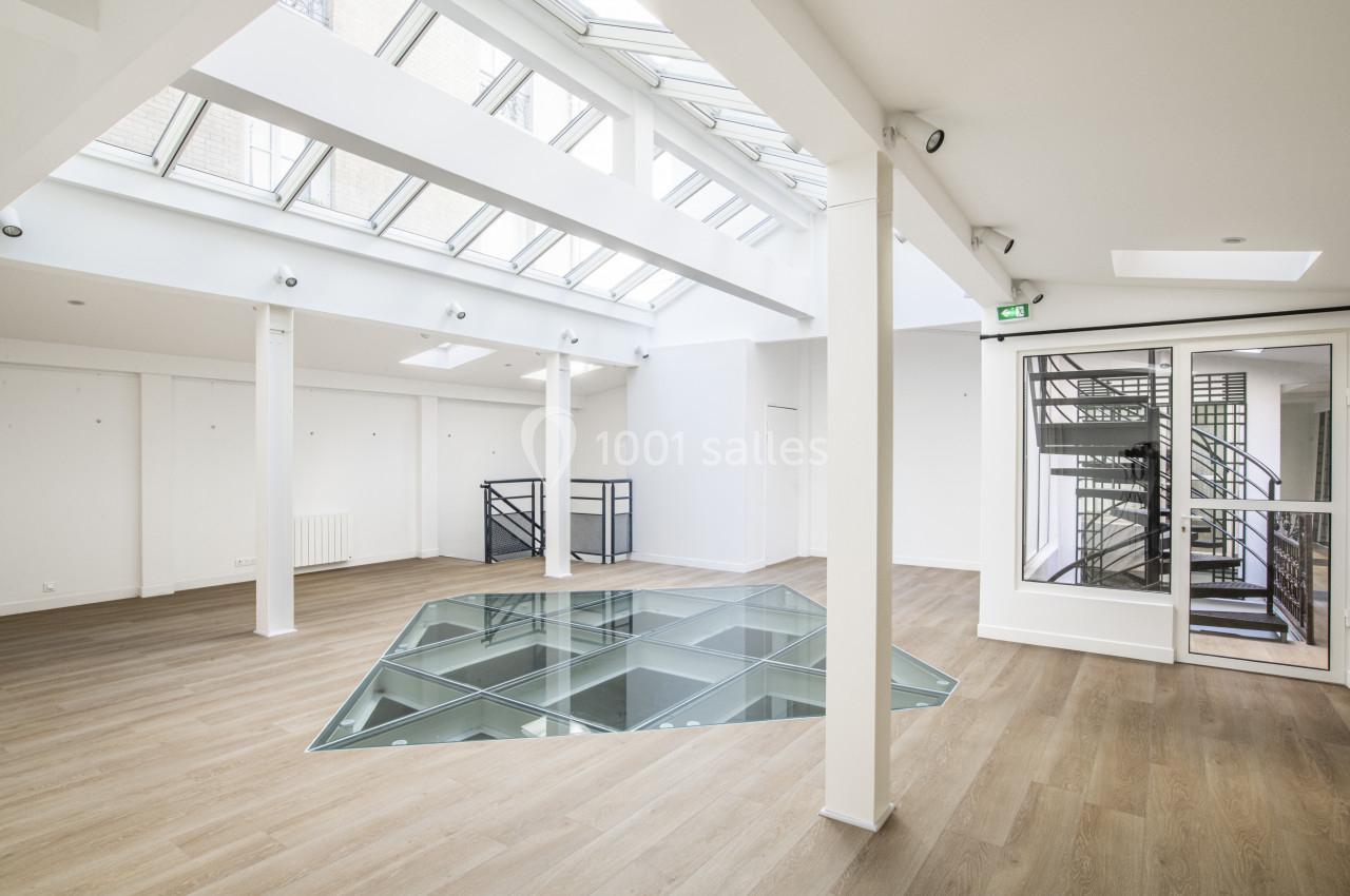 Salle lumineuse avec parquet, verrière au plafond, sol en verre et escalier visible derrière une porte vitrée.