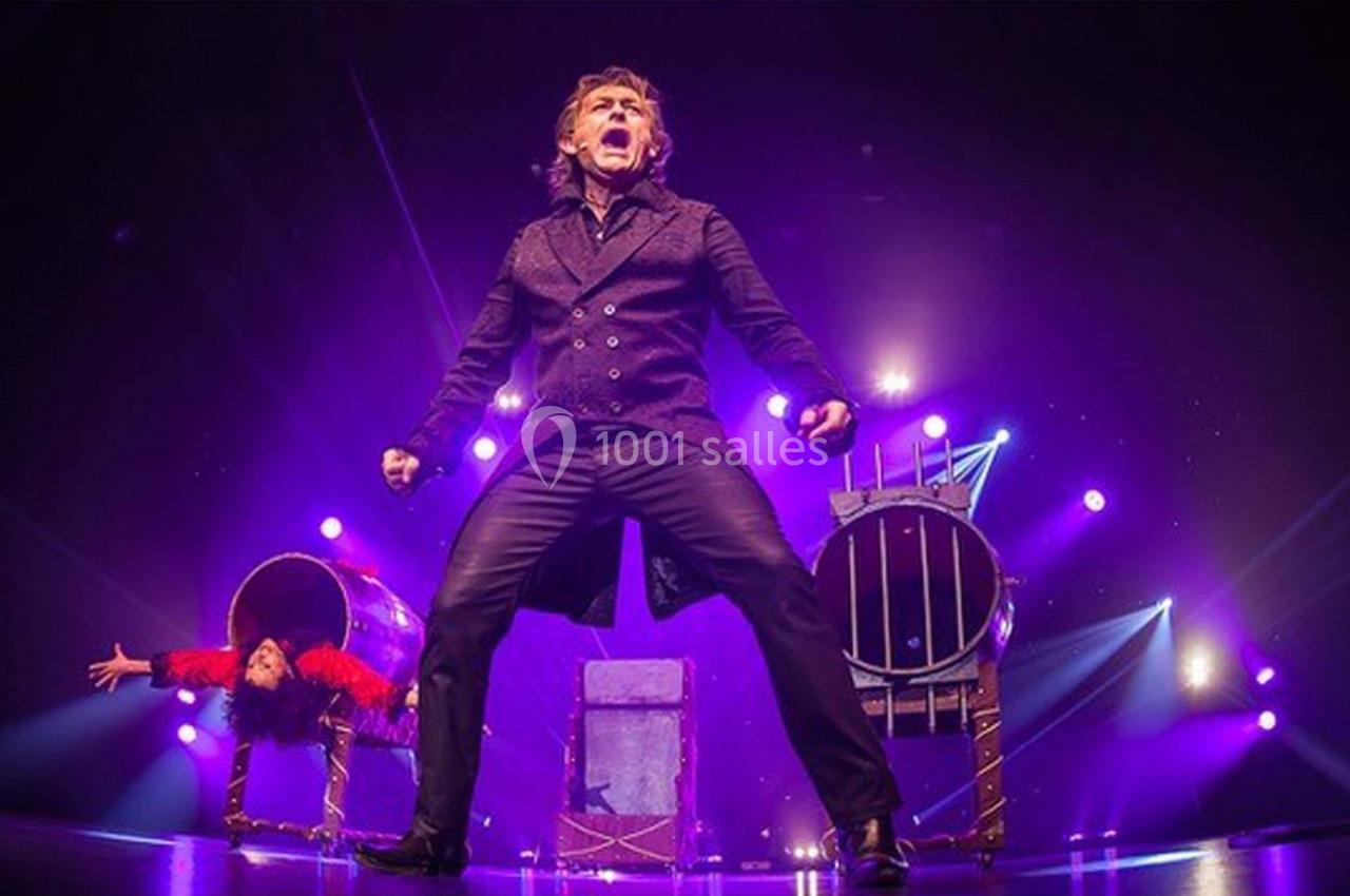 Un magicien sur scène effectue un numéro avec une assistante sortant d'une boîte, éclairé par des projecteurs violets.