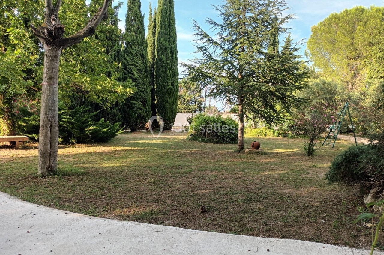 Jardin arboré avec pelouse, allée en béton, arbres variés et balançoire visible en arrière-plan.