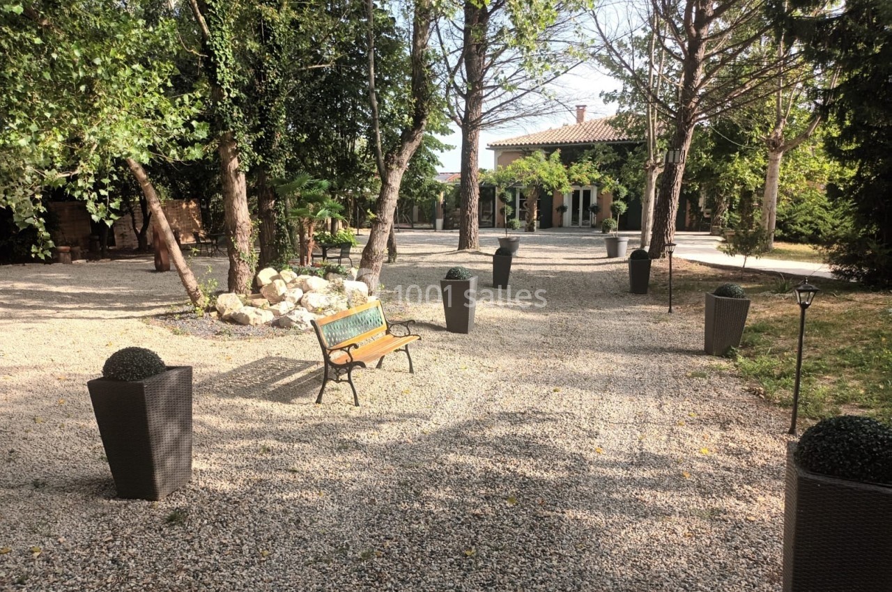 Allée gravillonnée bordée d'arbres, avec des pots décoratifs et un banc en bois face à une maison.