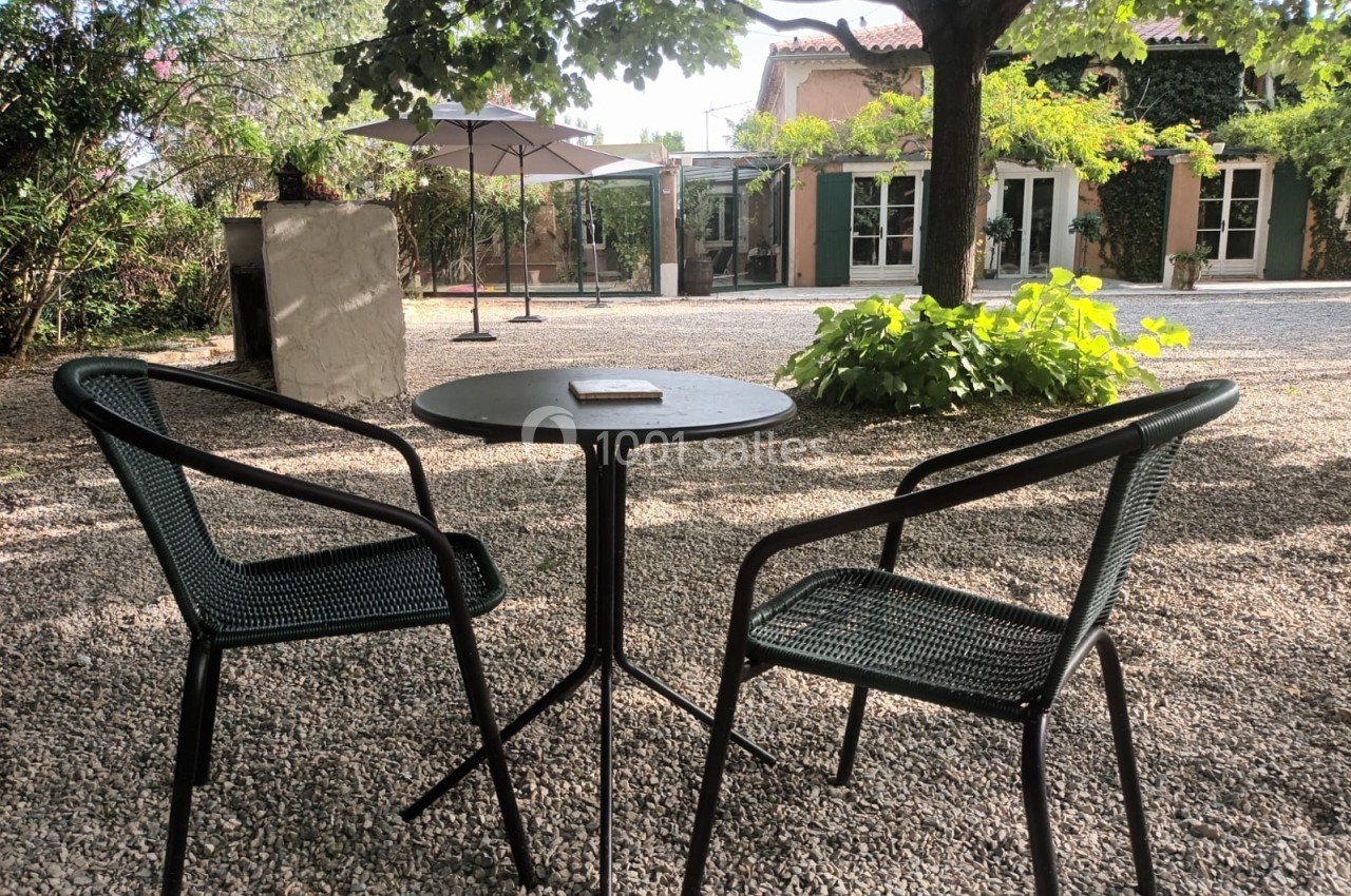 Deux chaises et une table ronde en métal noir sur un sol gravillonné, dans une cour ombragée par des arbres.