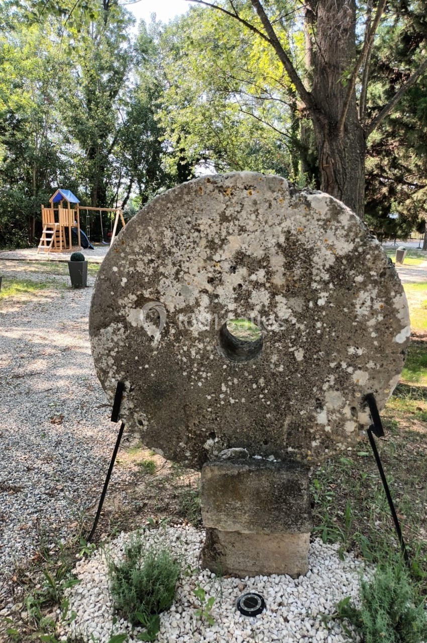 Meule en pierre ancienne exposée dans un jardin, entourée de gravier, avec des arbres et une aire de jeux en arrière-plan.