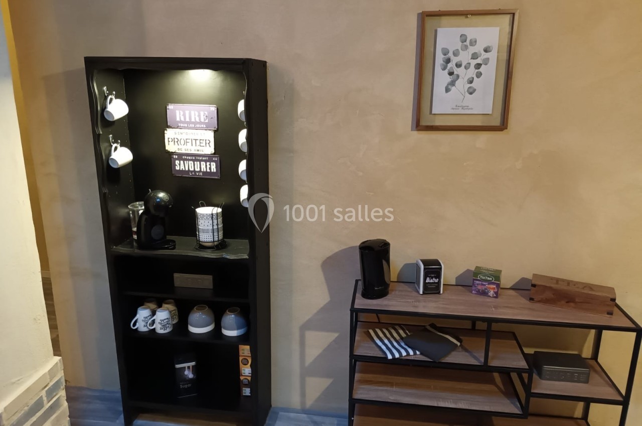 Meuble avec machine à café, tasses et accessoires, placé près d'une étagère et d'un mur décoré d'un cadre.
