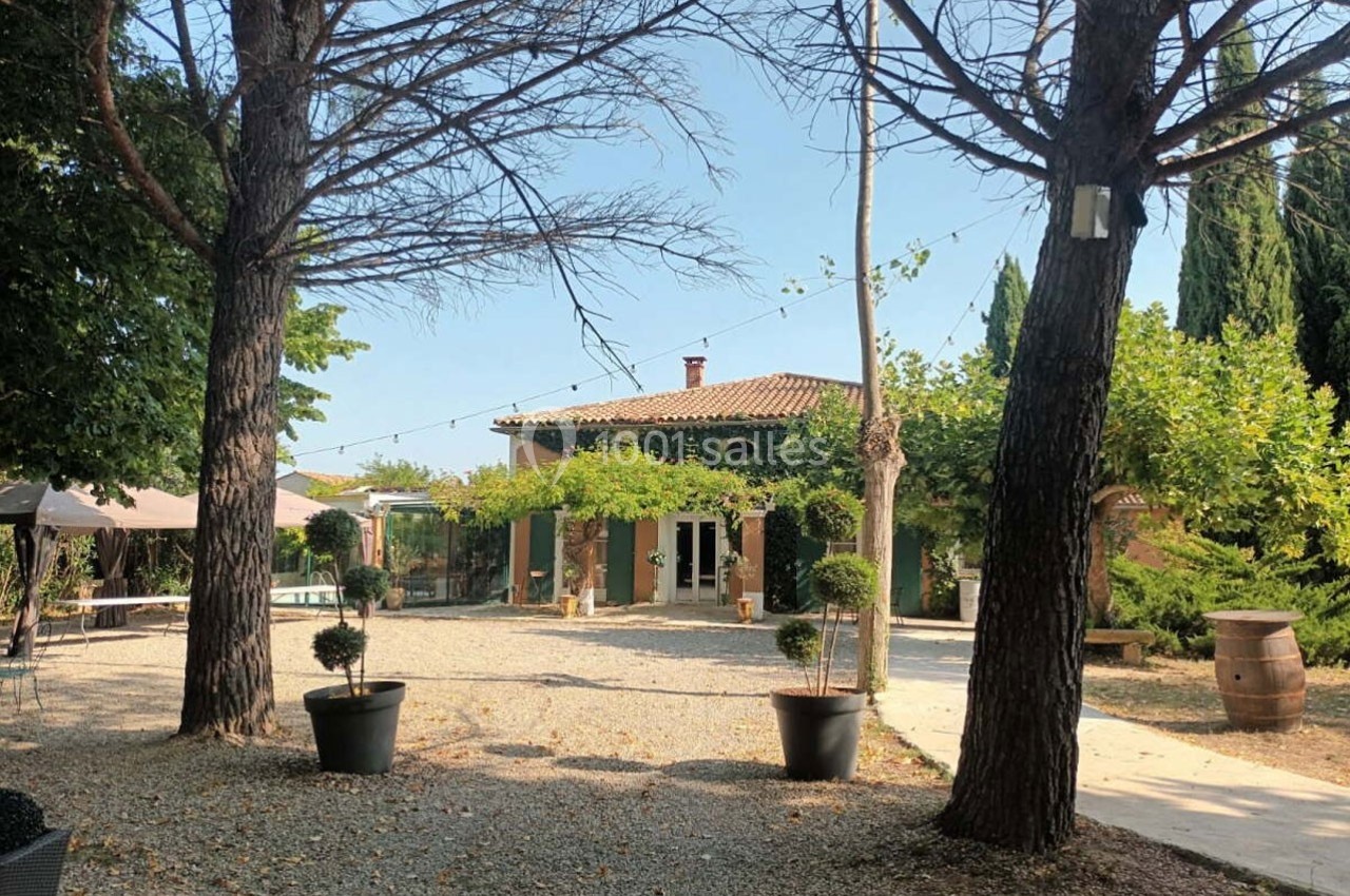 Maison provençale entourée d'arbres, avec une cour en gravier, des plantes en pot et une ambiance ensoleillée.
