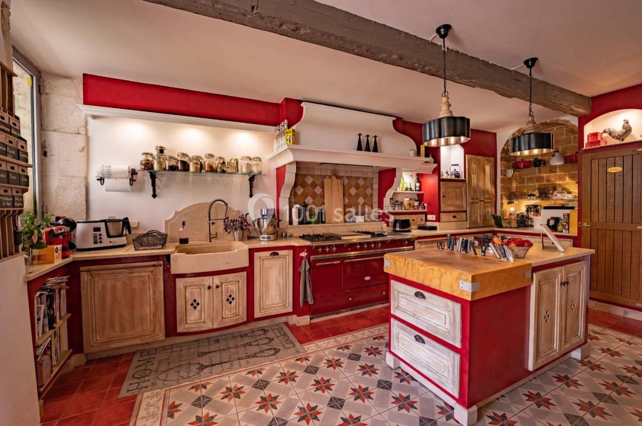 Cuisine rustique avec meubles en bois rouge et beige, plan de travail central, étagères ouvertes et carrelage décoratif.