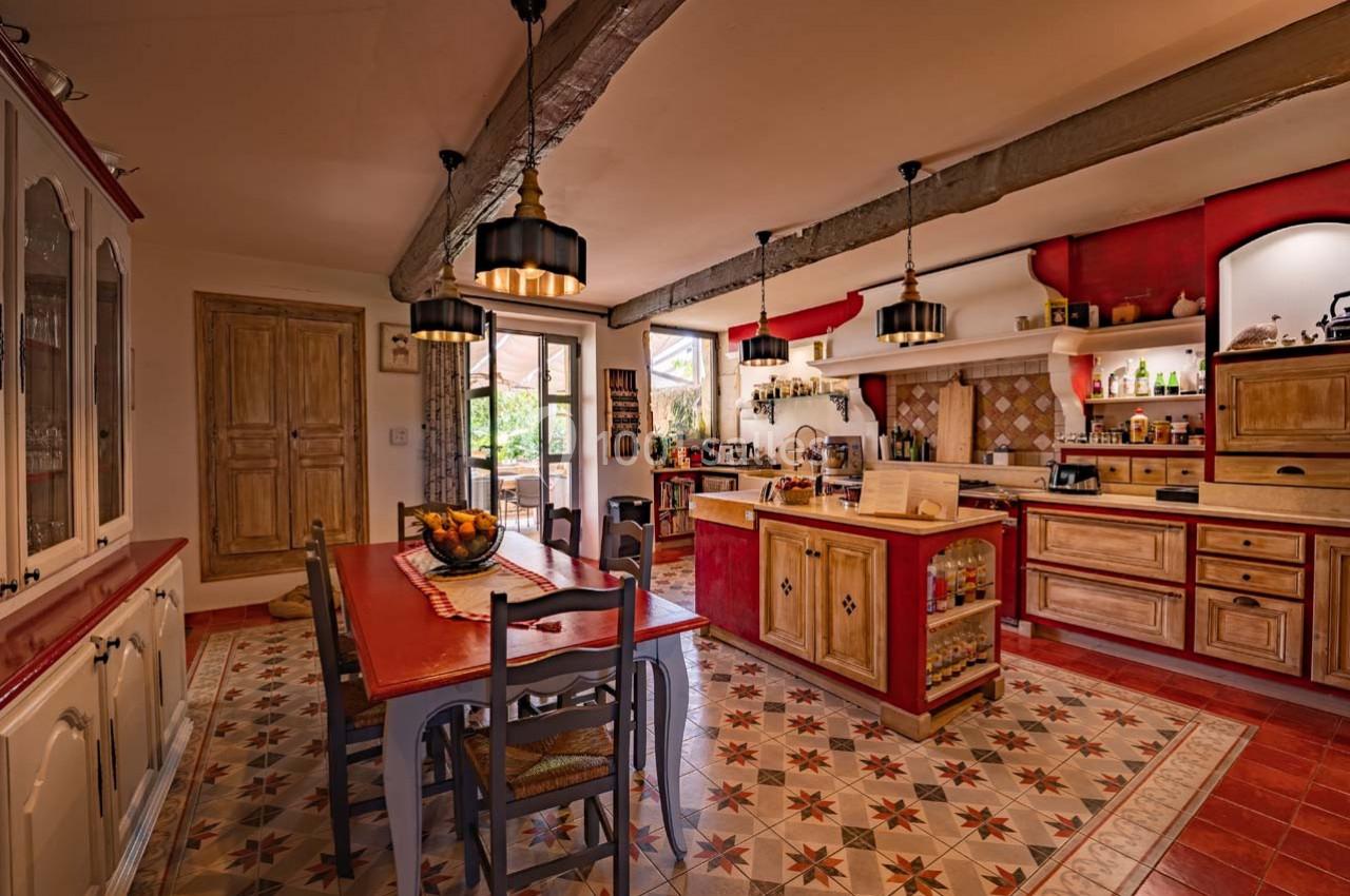 Cuisine rustique avec meubles en bois, carrelage à motifs, table à manger et lumière naturelle entrant par une porte-fenêtre.