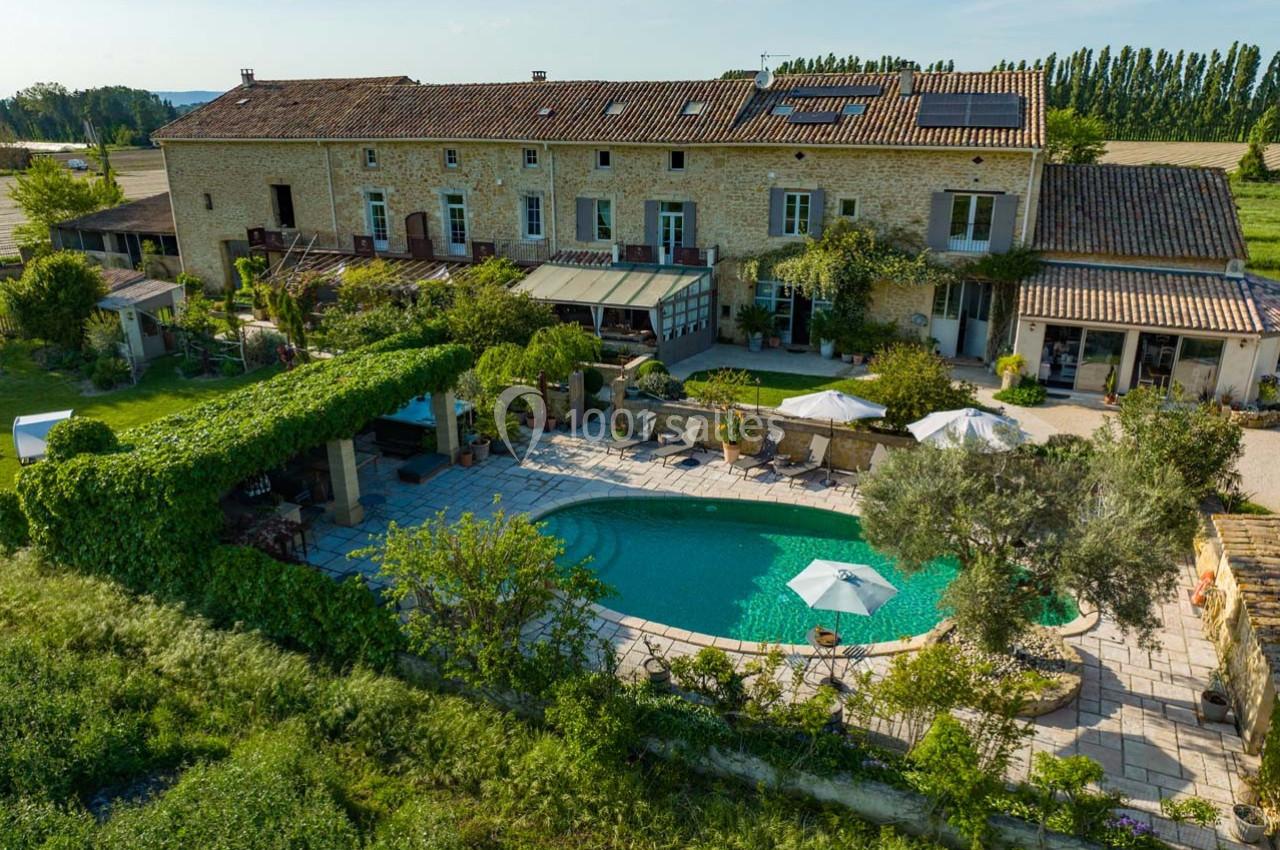 Vue aérienne d'une grande maison en pierre avec piscine, terrasse ombragée et jardin, entourée de champs verdoyants.