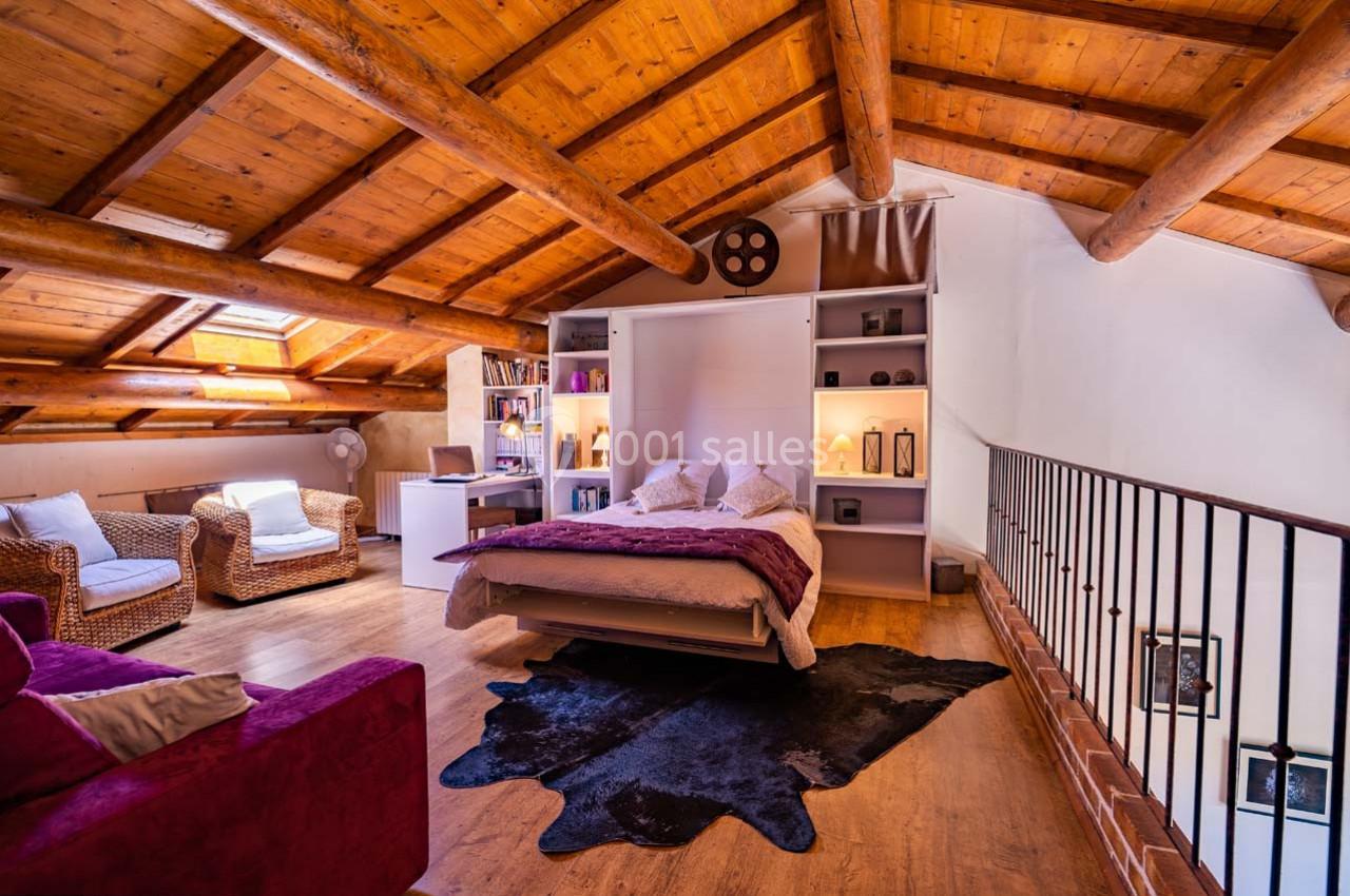 Chambre mansardée avec plafond en bois, lit central, fauteuils en osier, étagères et lumière naturelle.