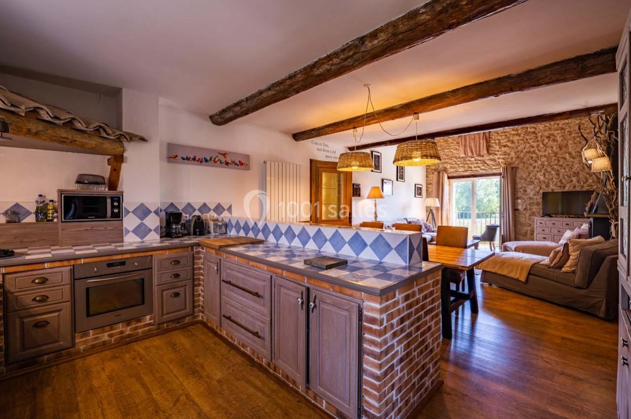 Cuisine ouverte rustique avec poutres apparentes, carrelage bleu, meubles en bois et vue sur un salon lumineux.
