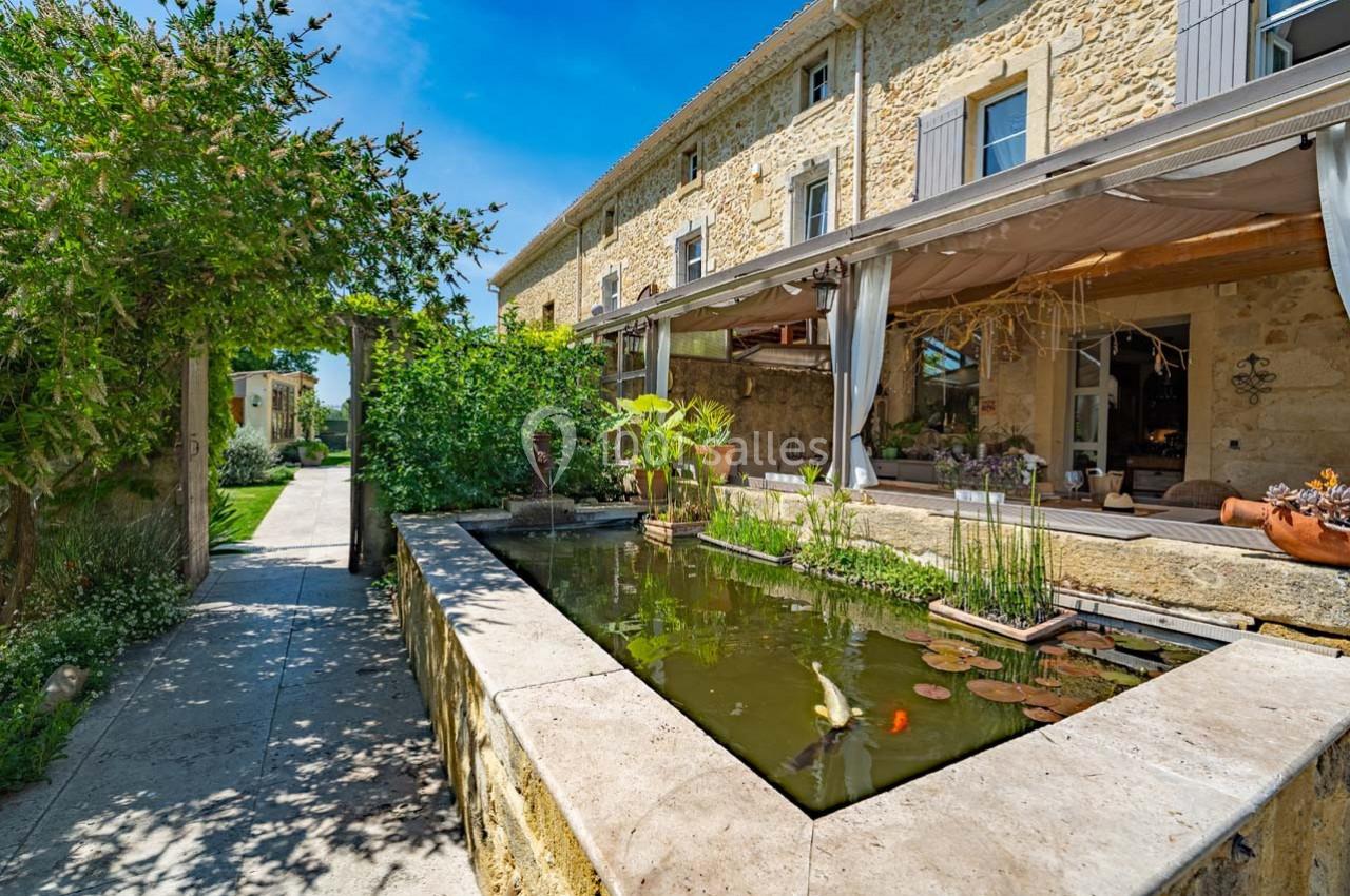 Bassin avec poissons et plantes aquatiques devant une maison en pierre avec terrasse couverte, entourée de verdure.
