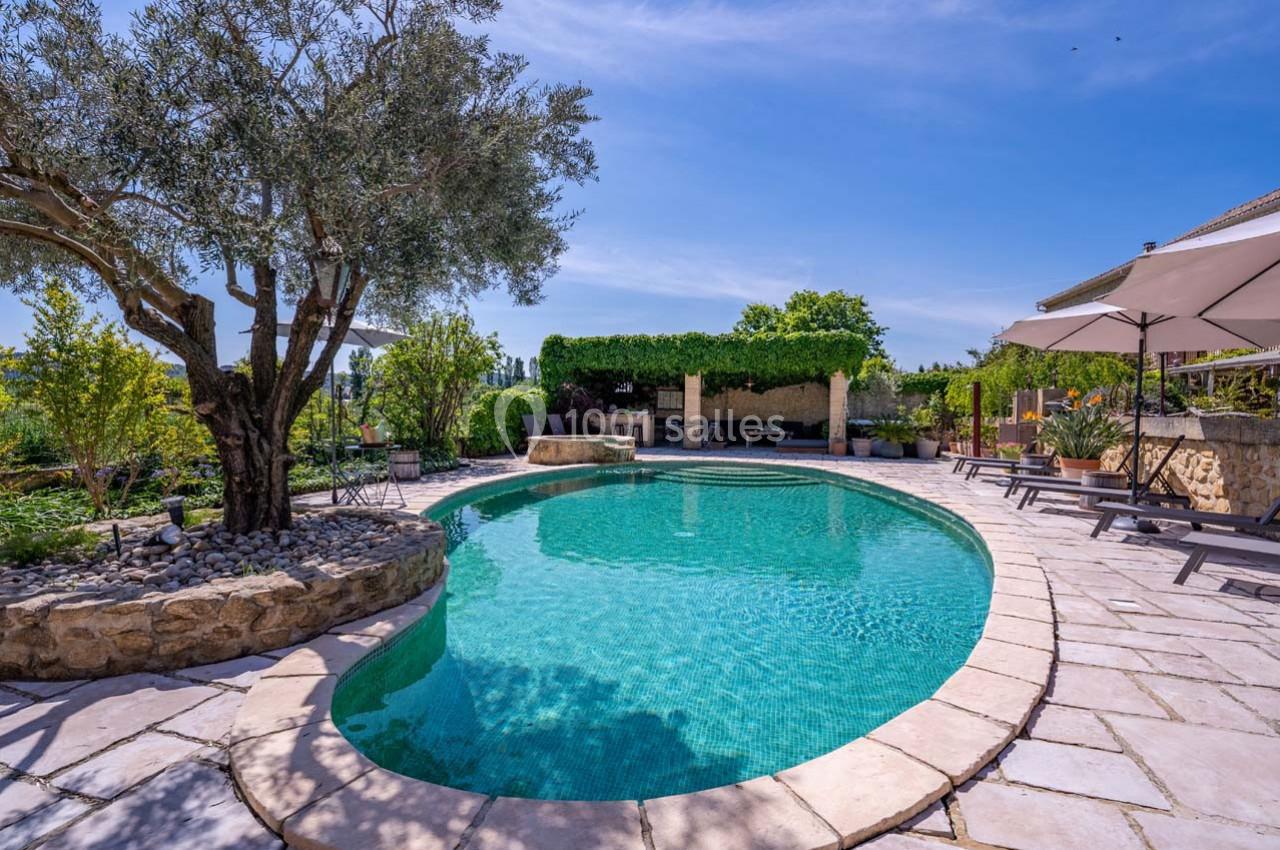 Piscine extérieure entourée de végétation, avec terrasse en pierre, chaises longues et parasols sous un ciel dégagé.