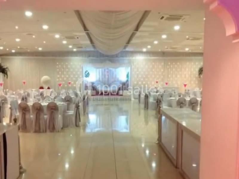 Salle de réception décorée avec des tables rondes, chaises drapées et éclairage tamisé, prête pour un événement.