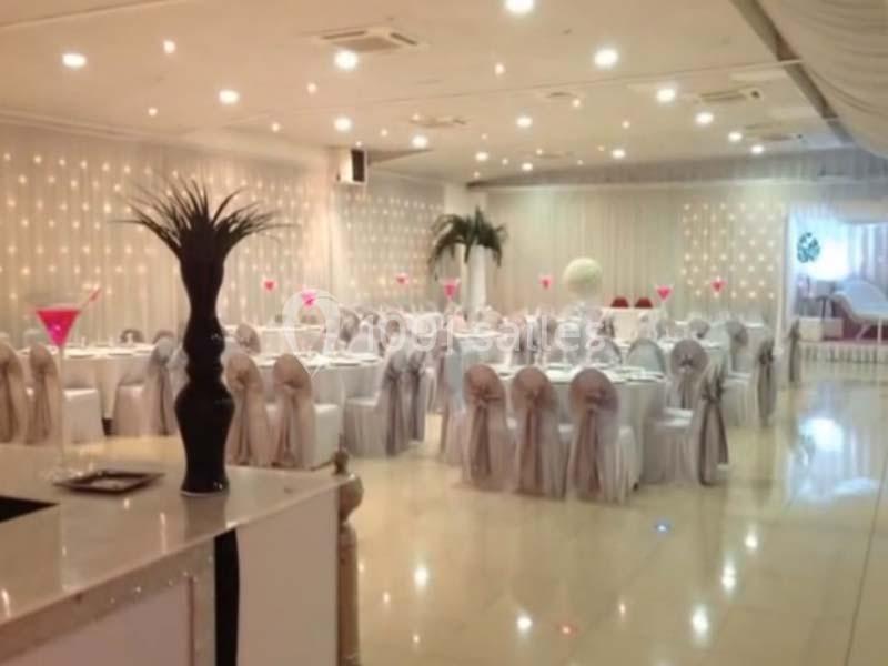 Salle de réception décorée avec des tables rondes, nappes blanches, chaises ornées de nœuds et éclairage tamisé.