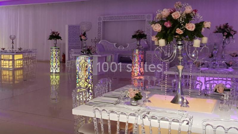 Salle de réception élégante avec tables décorées, chaises transparentes et arrangements floraux sur chandeliers argentés.