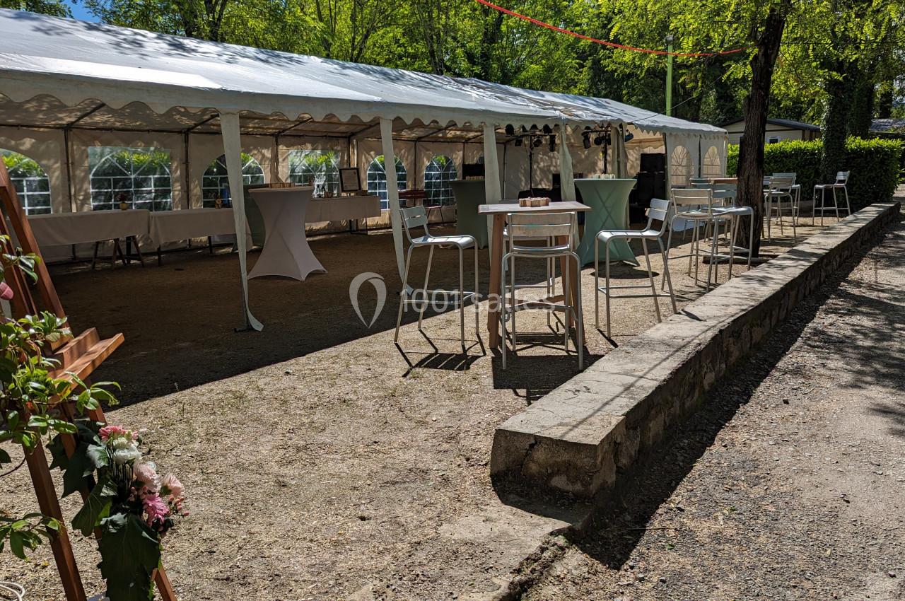 Espace extérieur avec une grande tente blanche, des tables hautes et des chaises, entouré d'arbres sous un ciel ensoleillé.