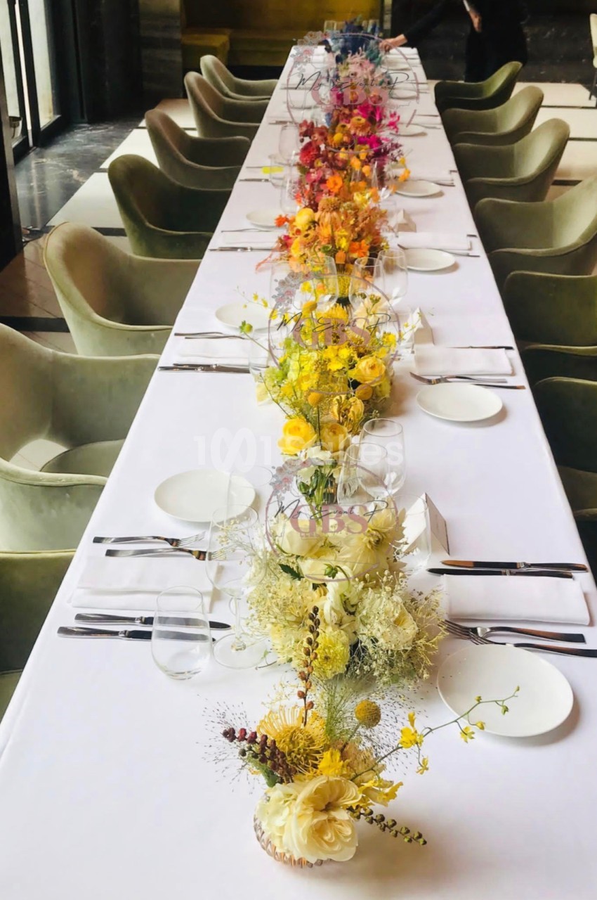 Table dressée avec une nappe blanche, des couverts élégants et une rangée de compositions florales colorées au centre.